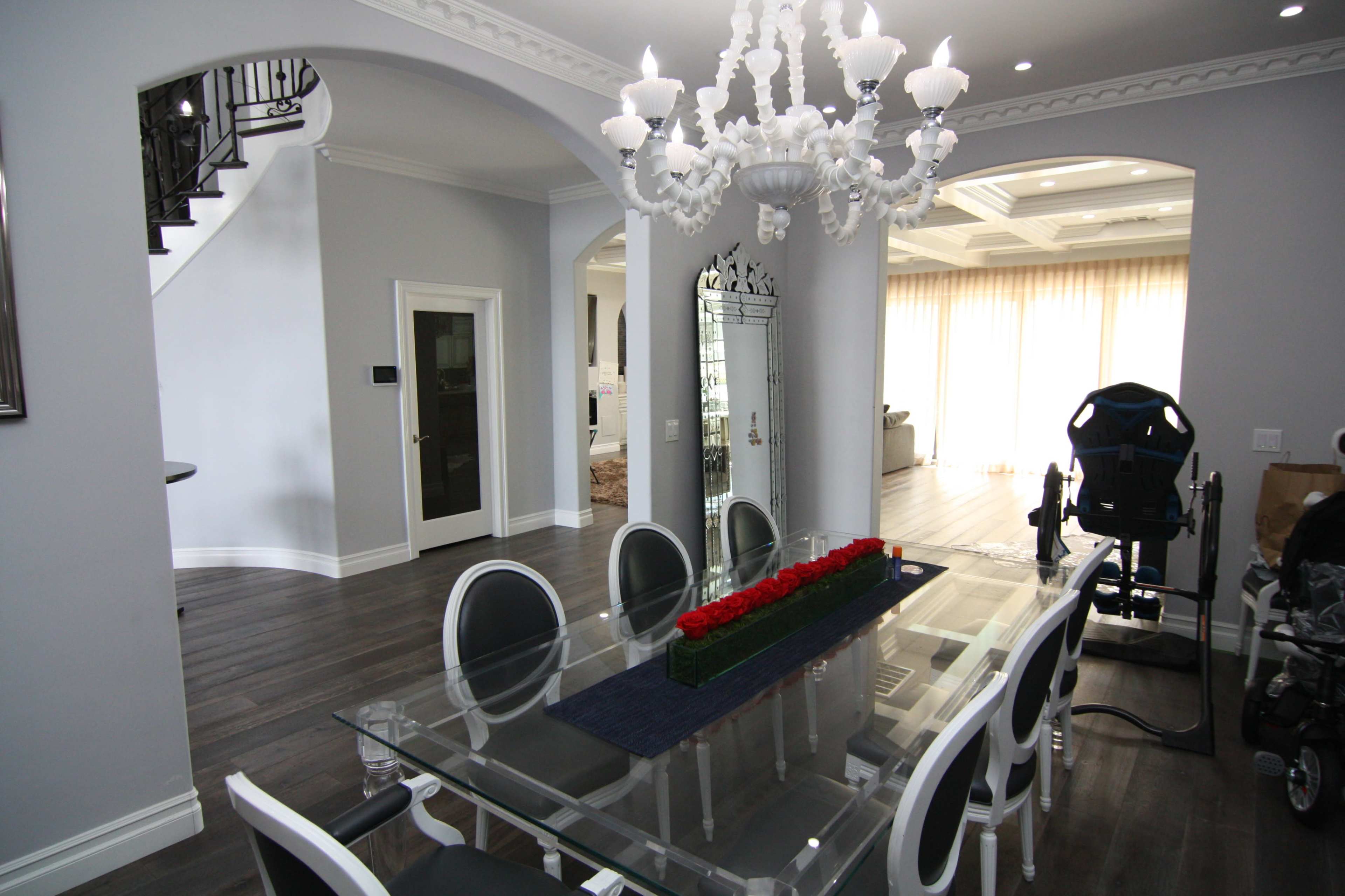 The image shows a modern dining area with a glass table, white chairs, and a chandelier, adjacent to a staircase and a bright living space.