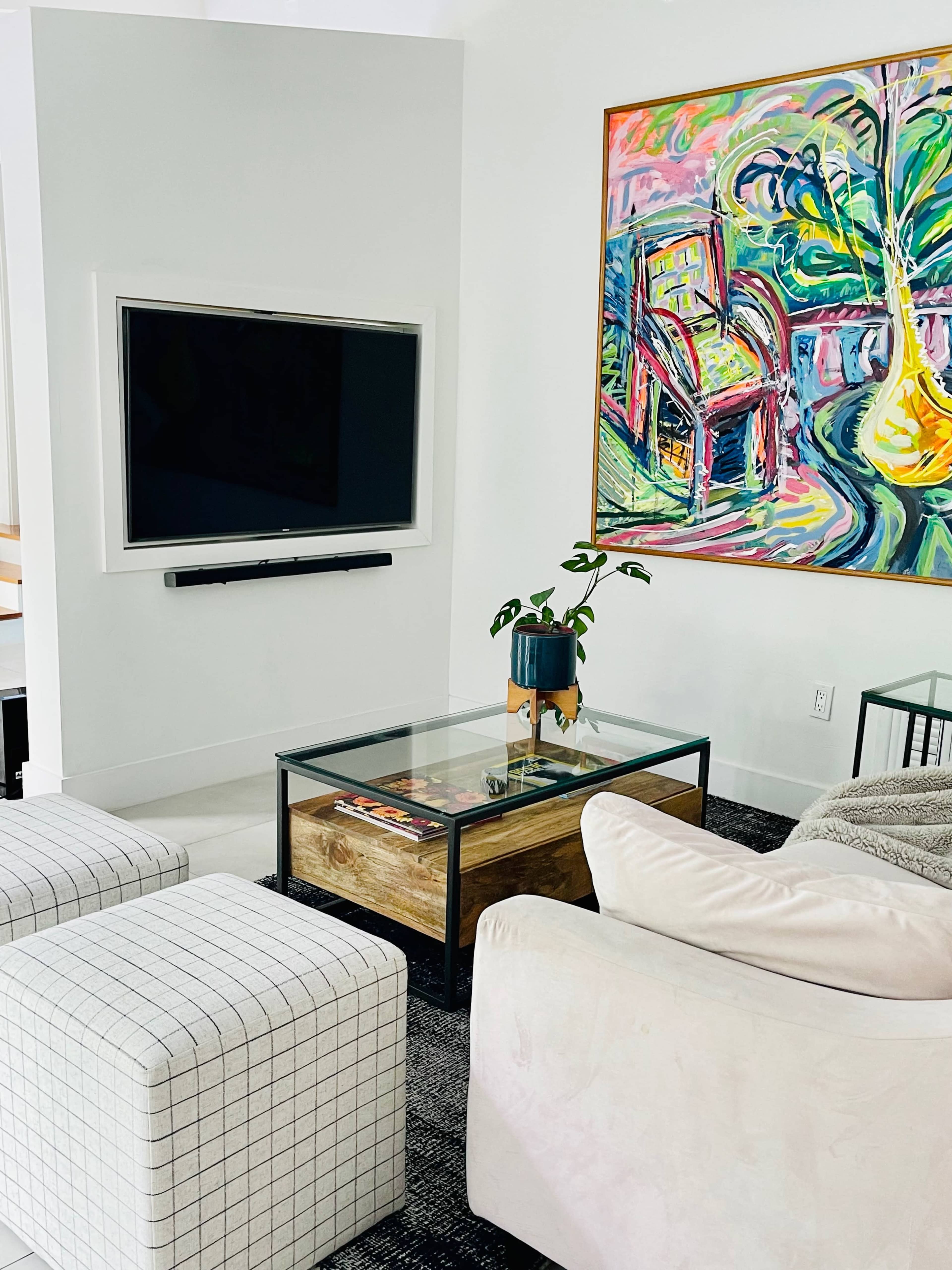 A modern living room with a television mounted on the wall, a glass coffee table, patterned ottomans, and a colorful abstract painting hanging nearby.
