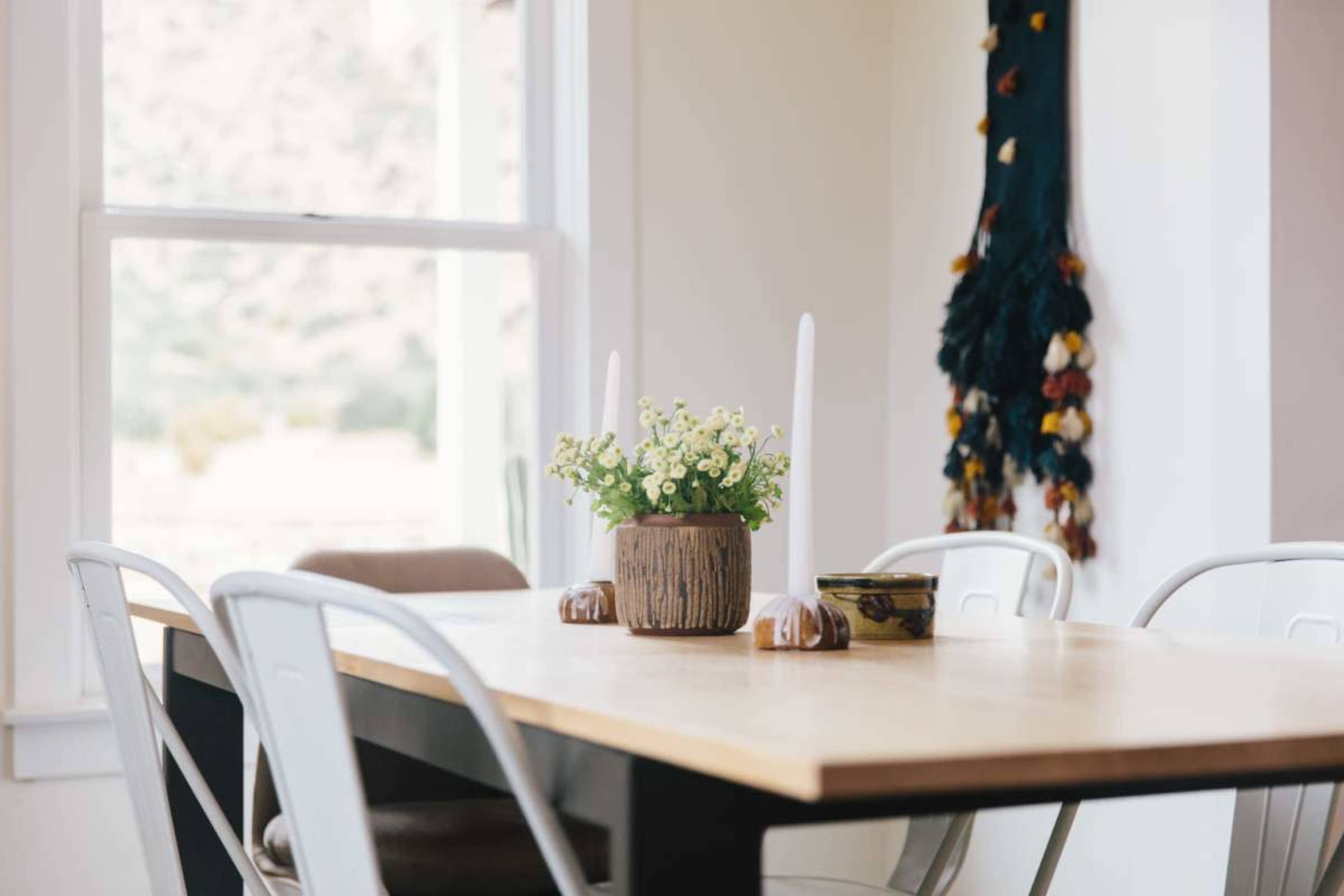 A wooden dining table is set with a floral centerpiece and two white candles, surrounded by metal chairs in a well-lit room.
