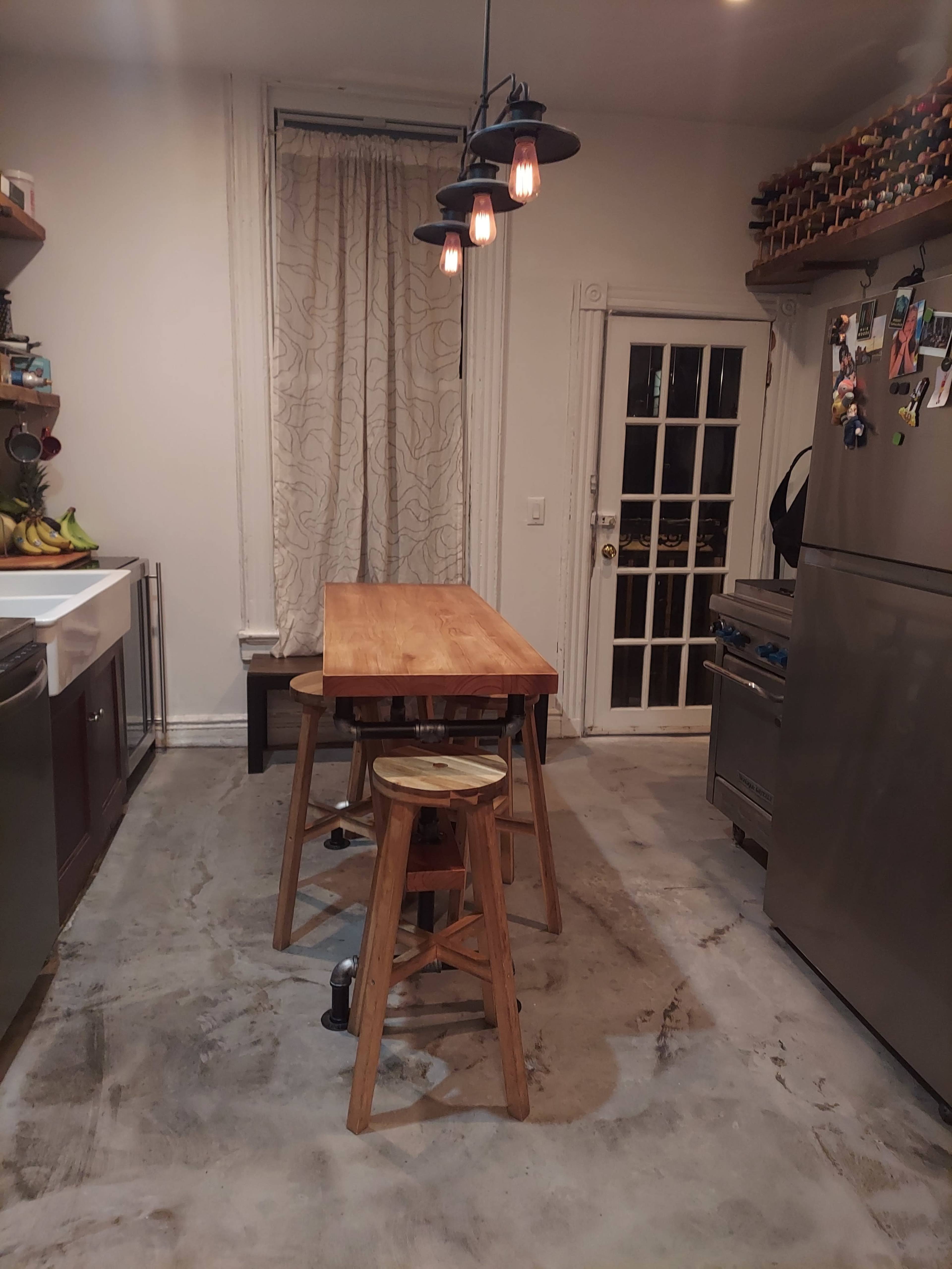 A wooden table with stools is positioned in a kitchen featuring a gray tiled floor and a single light fixture hanging above.