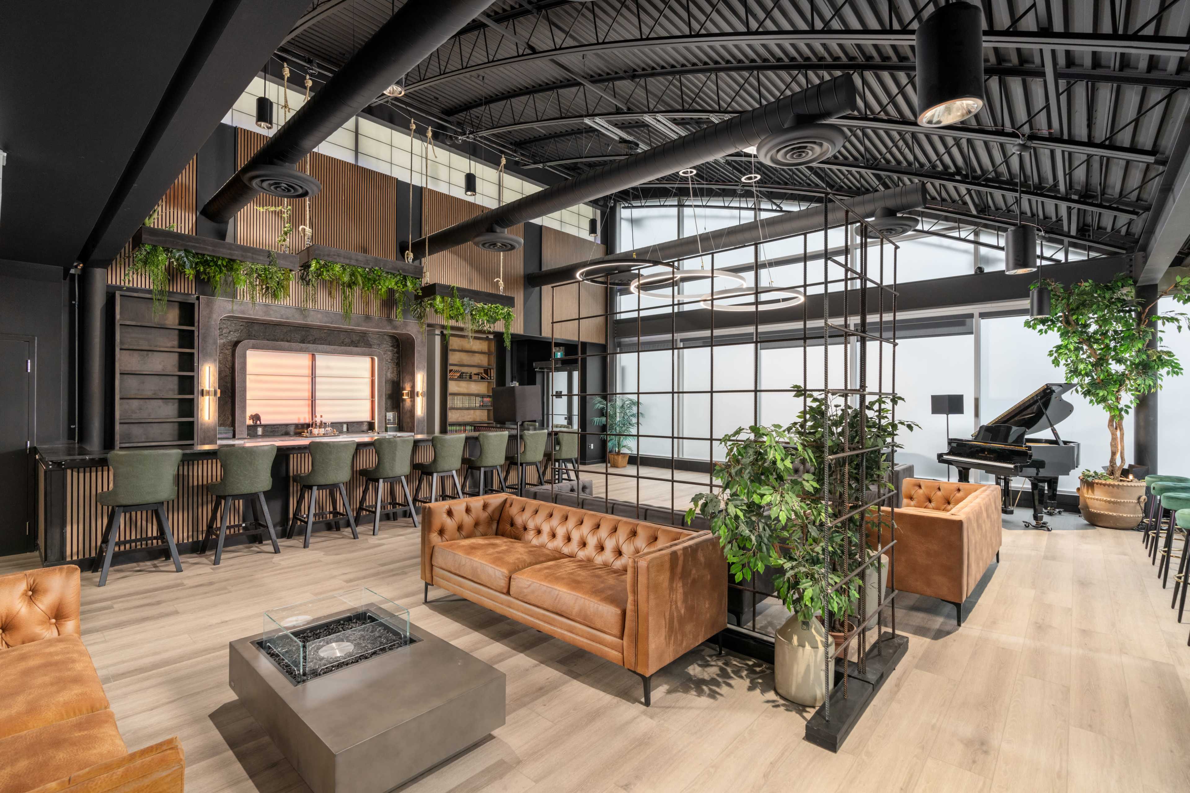 A modern lounge area featuring a bar with seating, a piano in the corner, and brown leather sofas arranged around a central fire pit.