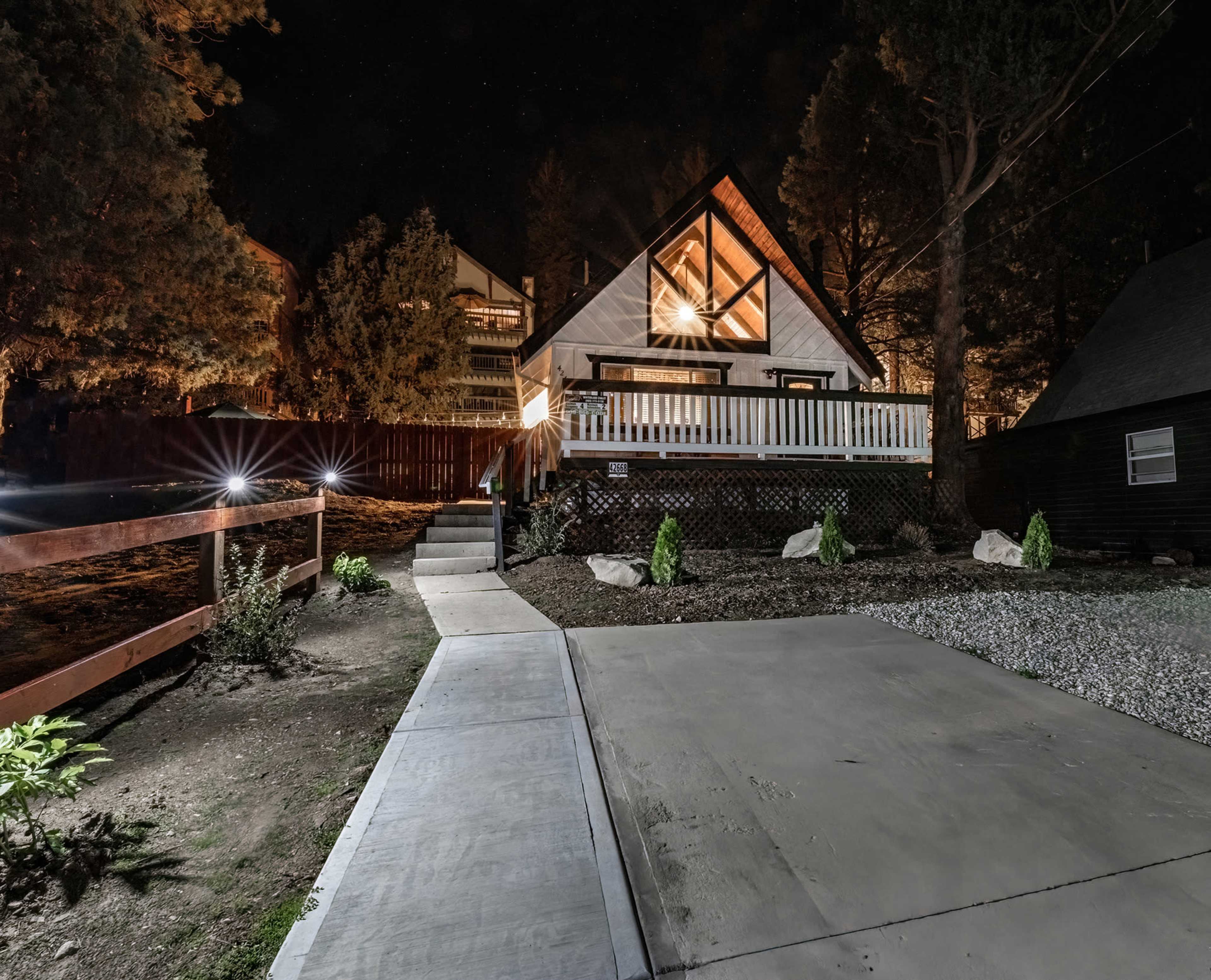 A modern A-frame house is illuminated at night, surrounded by trees and a landscaped yard with a paved walkway leading to it.