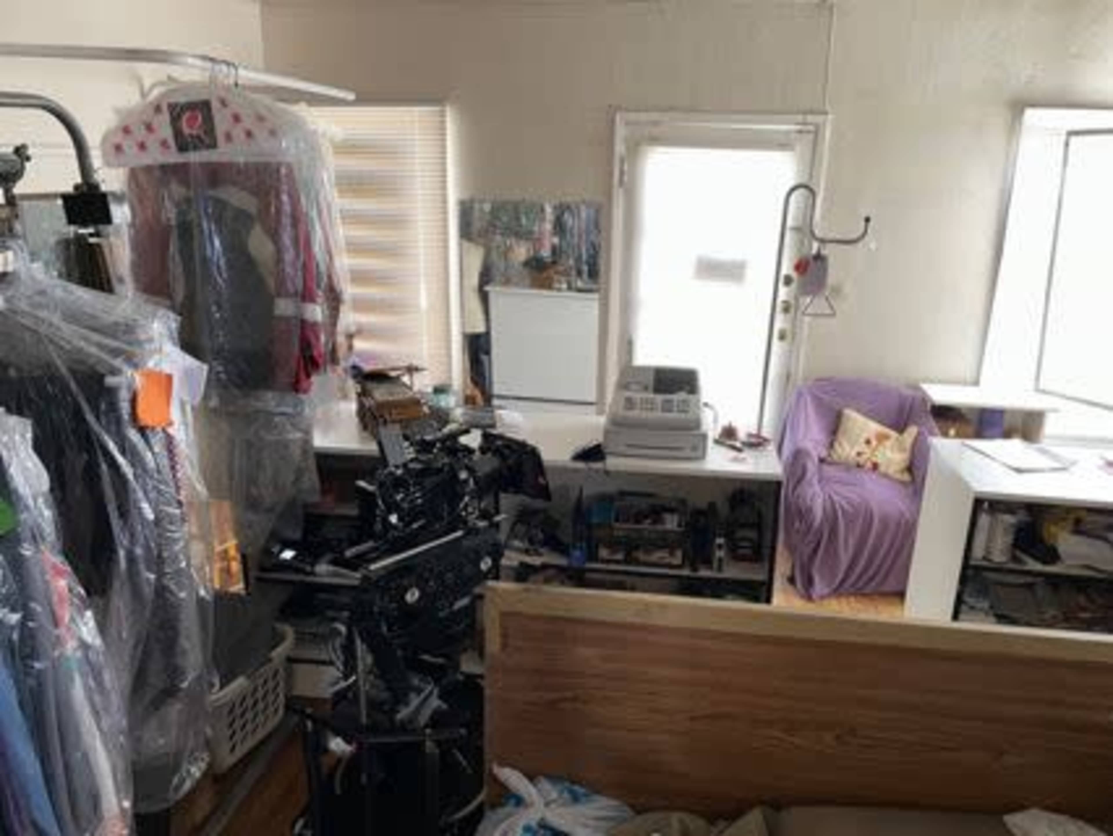 The image shows a cluttered room with various clothing items covered in plastic, a sewing machine, and a purple chair near a window.