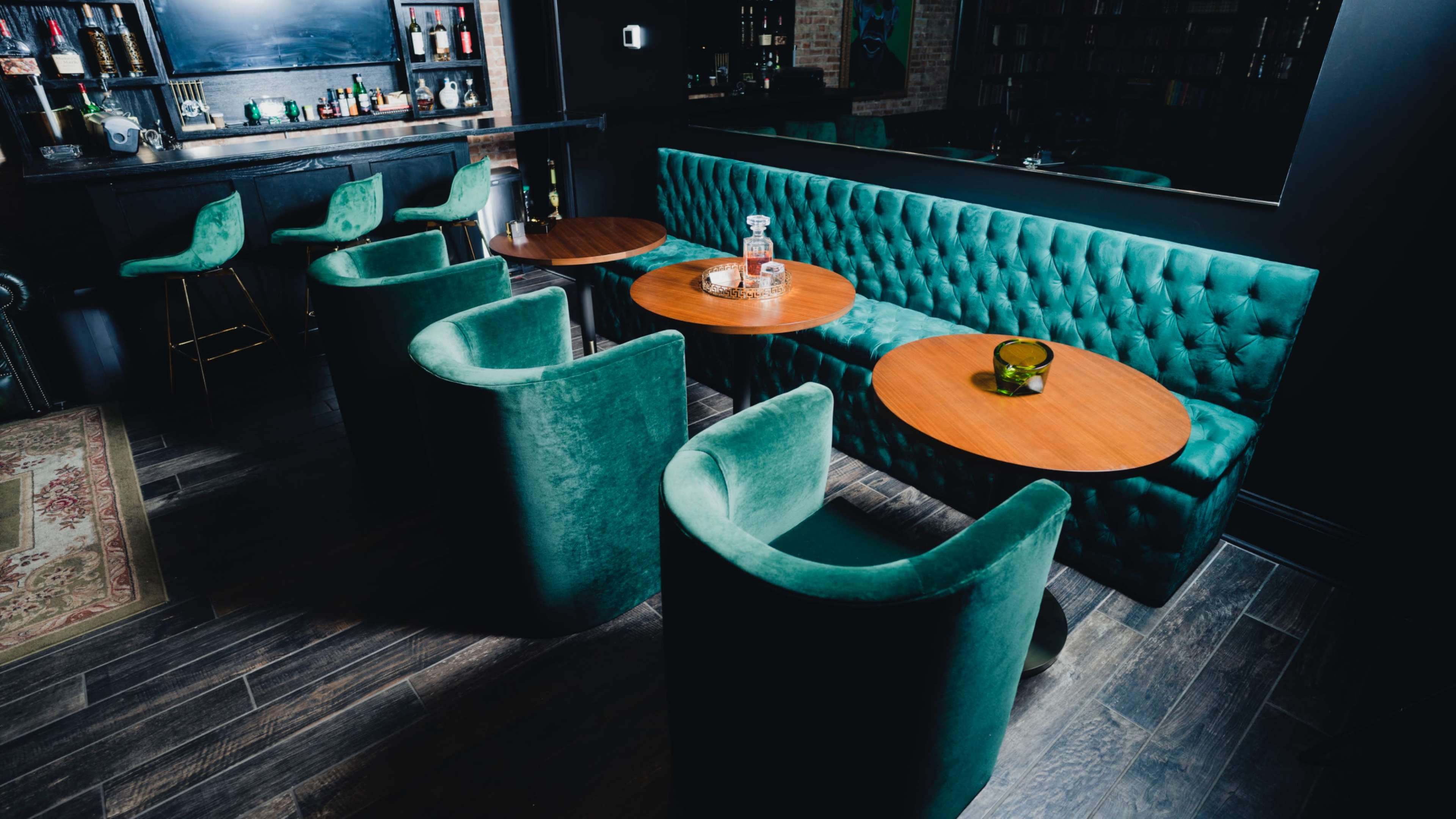 A dimly lit bar area featuring green upholstered seating, wooden tables, and a well-stocked bar in the background.