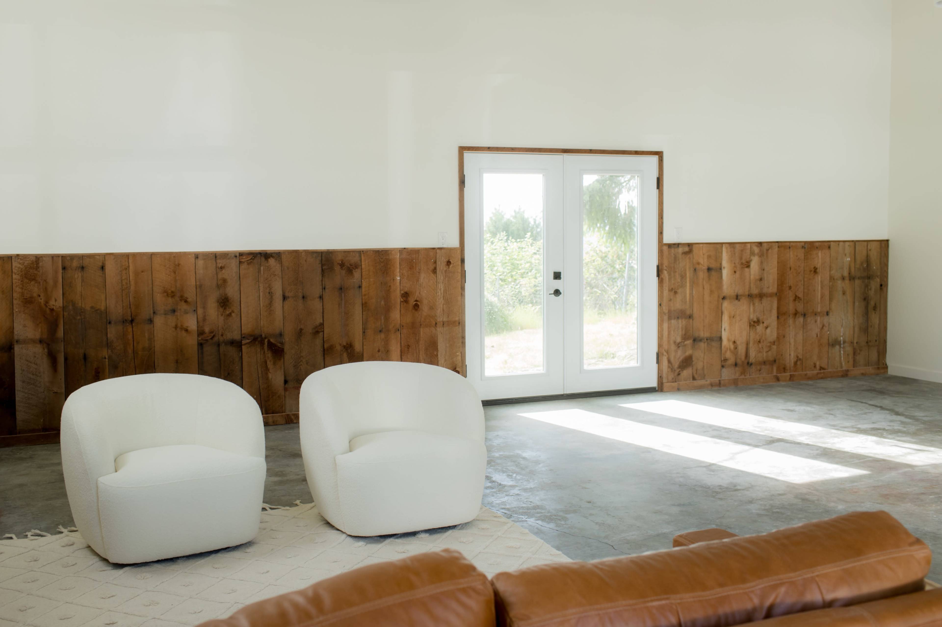 A pair of white chairs are positioned in a bright room with a wooden accent wall and a door leading to an exterior view.