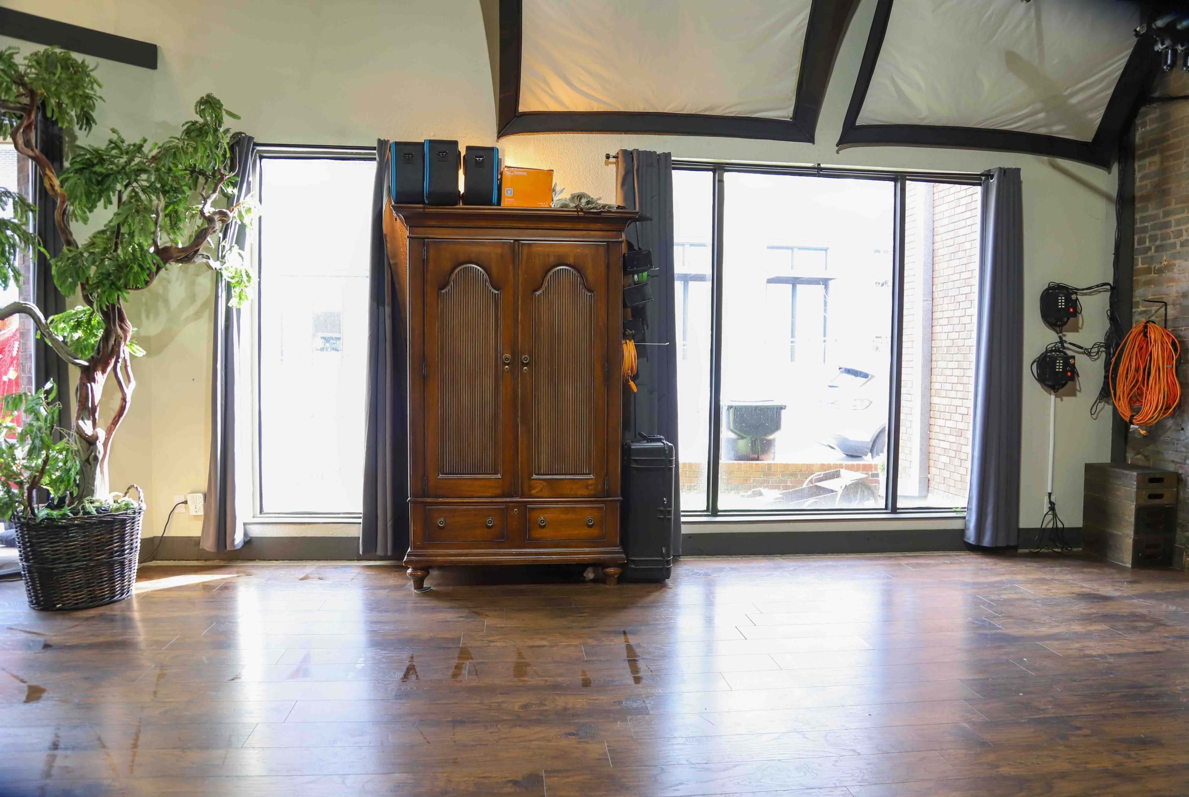 A spacious room with hardwood flooring, featuring a wooden wardrobe against a wall, large windows letting in natural light, and various equipment visible in the background.