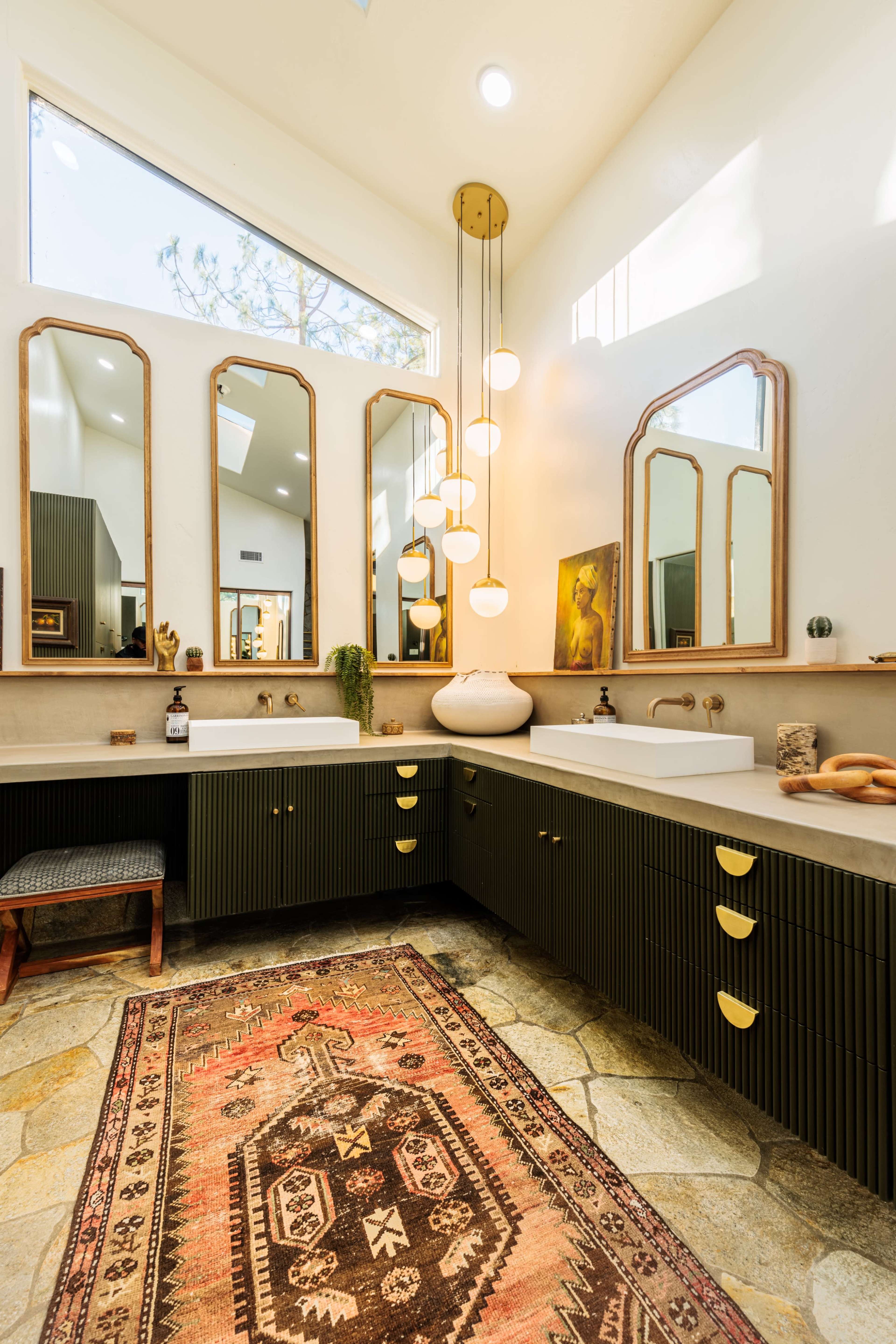 The bathroom features a double vanity with green cabinets, multiple mirrors, a light fixture hanging above, and a patterned rug on a stone floor.