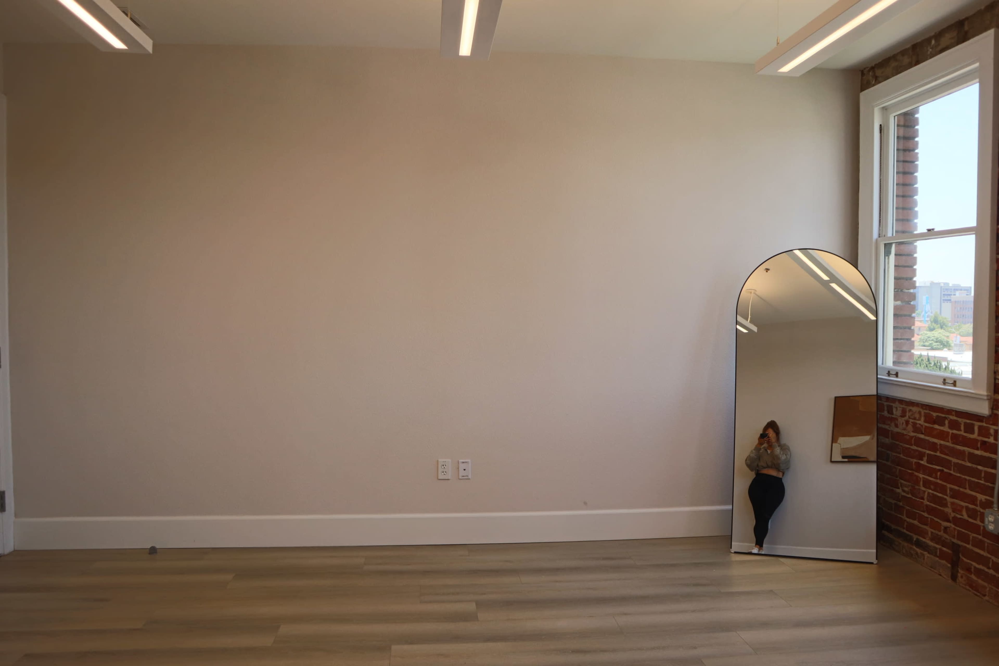 A spacious, empty room features large windows, a wooden floor, and a tall, arched mirror against the wall.