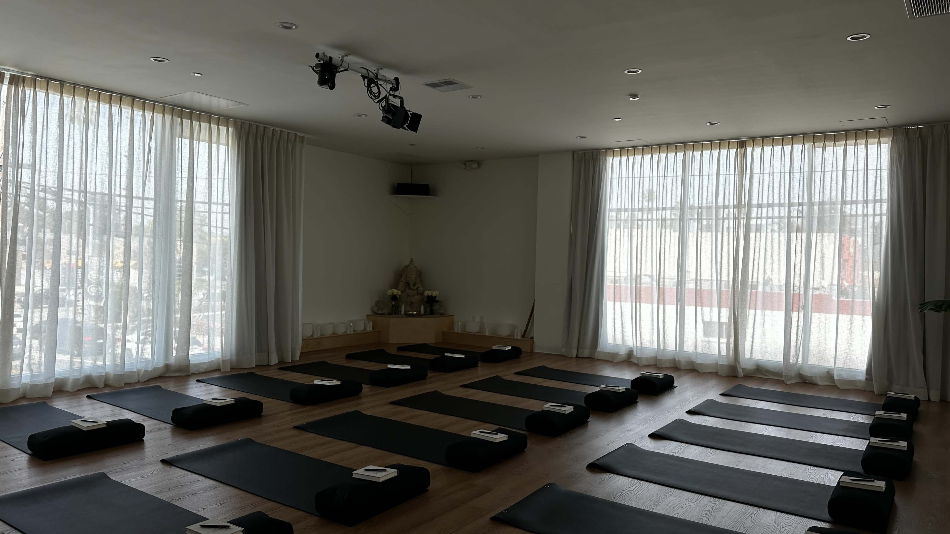 The image shows a yoga studio with neatly arranged mats on the wooden floor and large windows with sheer curtains letting in natural light.