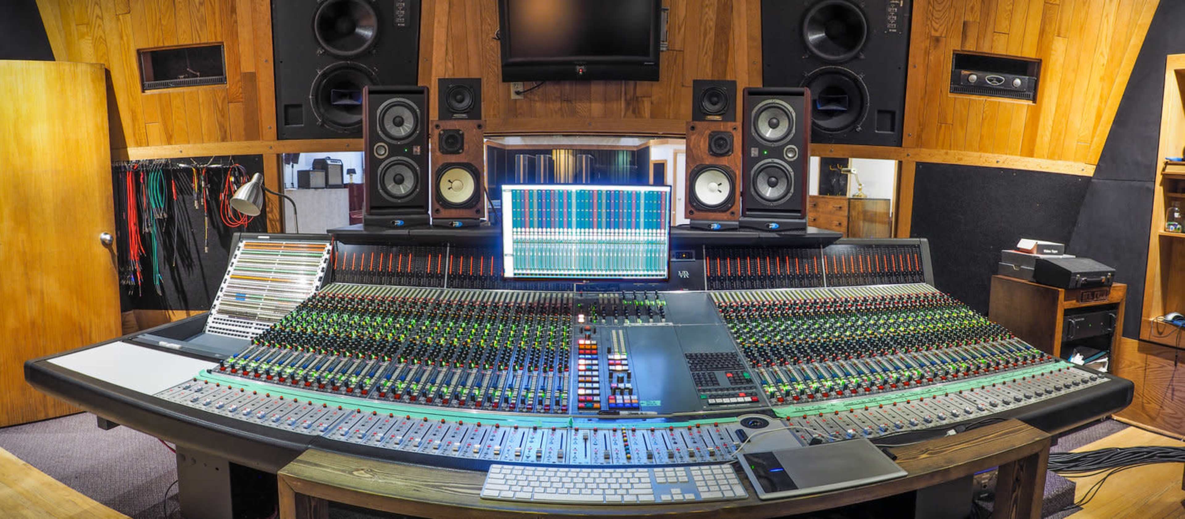 A panoramic view of a professional music studio with a large mixing console, computer monitor, and high-quality speakers.