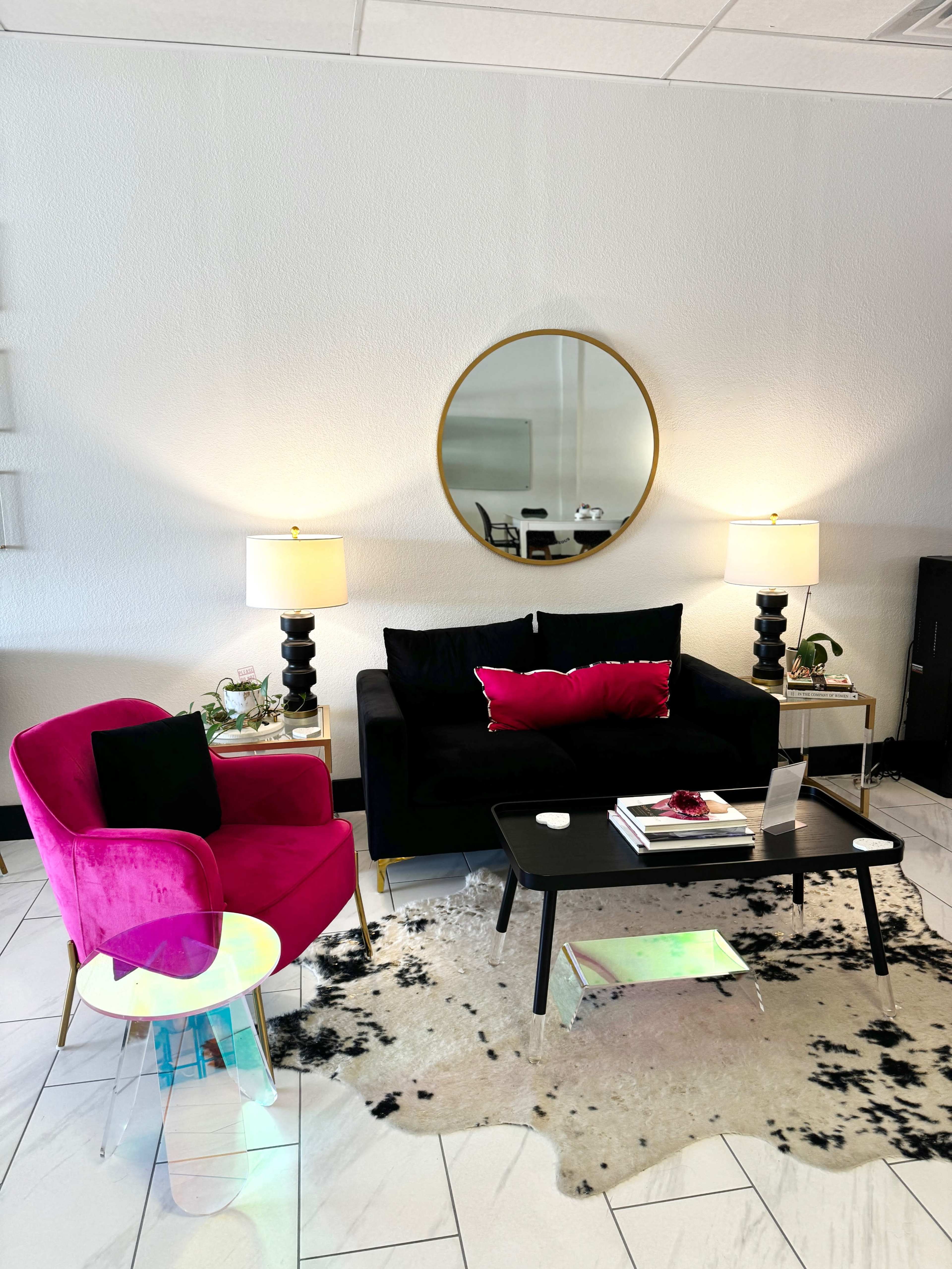A modern living room featuring a pink armchair, a black sofa with decorative pillows, a coffee table, and a patterned area rug, all softly illuminated by two table lamps.