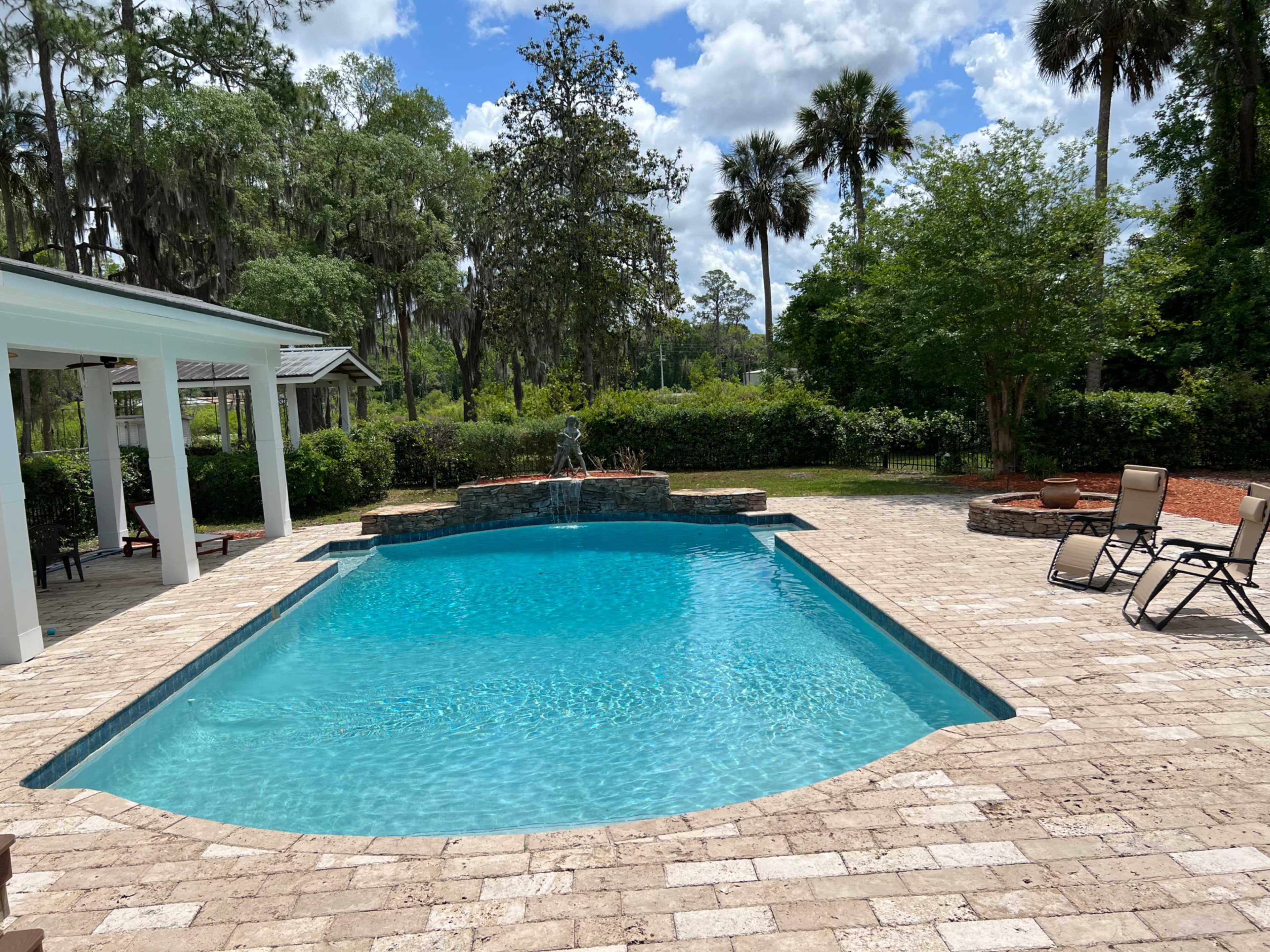A clear blue swimming pool surrounded by a paved patio and lush greenery, with a water feature and palm trees in the background.