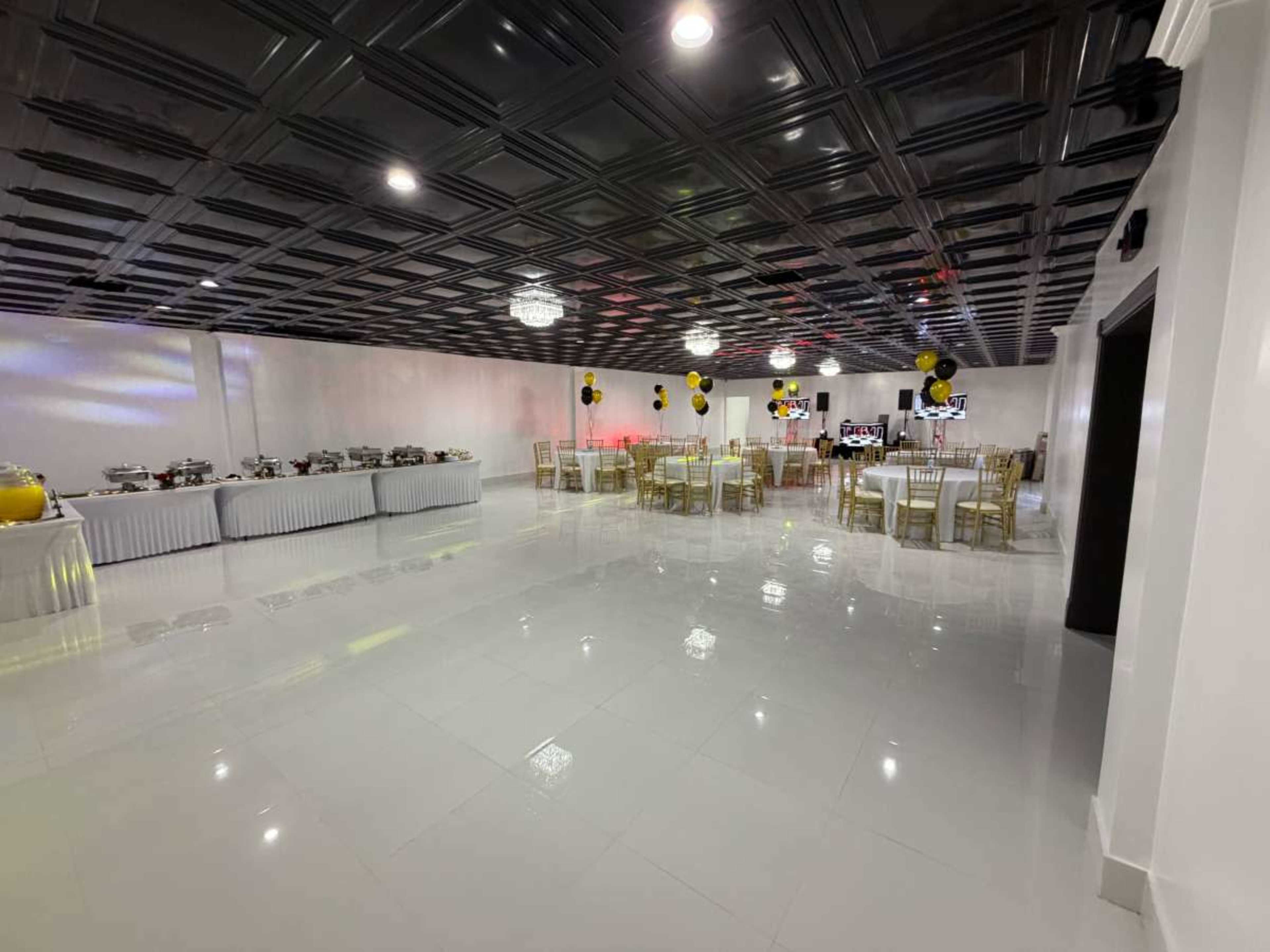 The image shows a spacious event hall with white walls, a black ceiling, and tables set for dining, along with a buffet setup along one wall and decorative balloons.