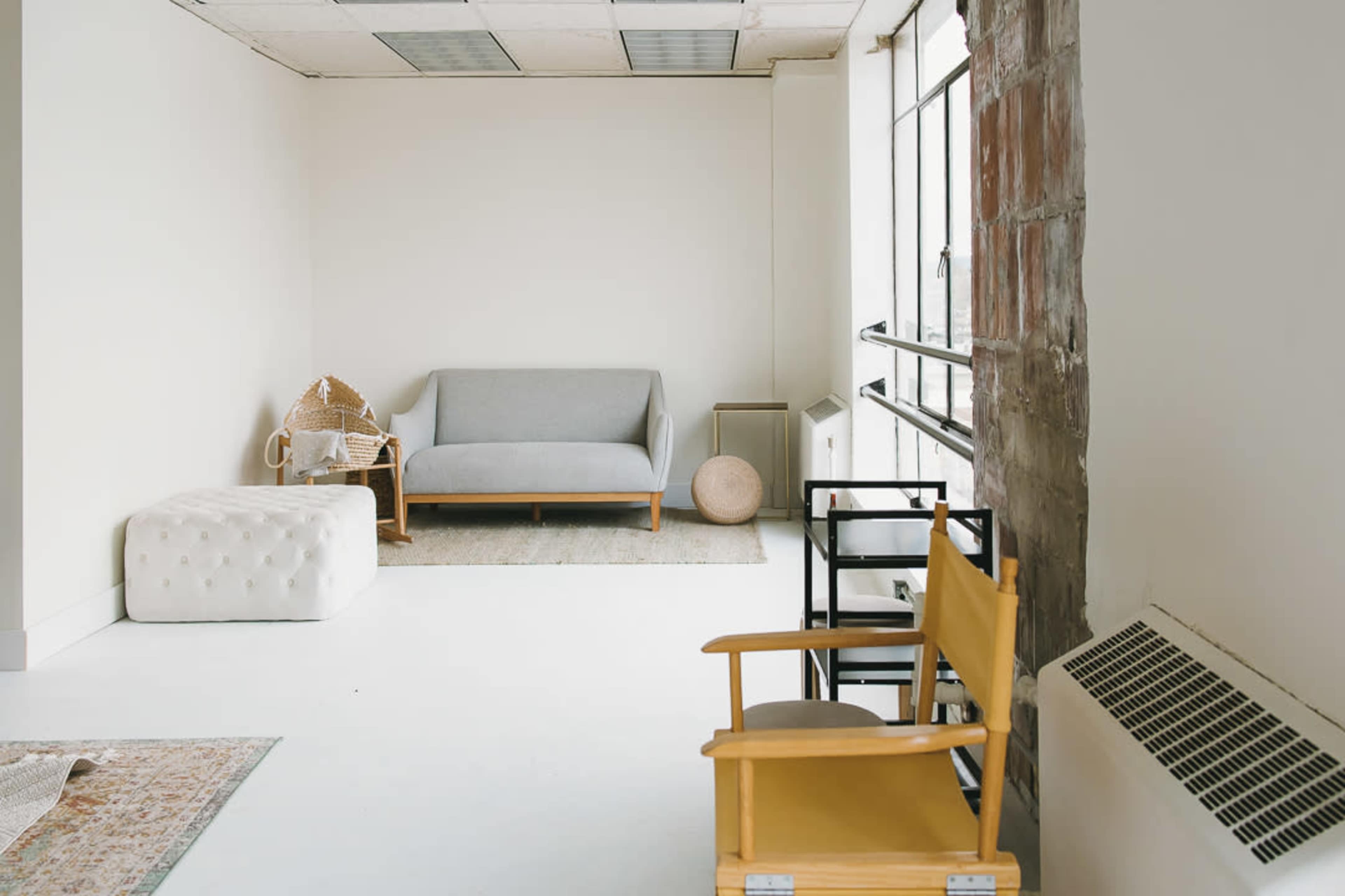 The image shows a minimalist room featuring a gray sofa, a white pouf, a wooden chair, and large windows that allow natural light to fill the space.