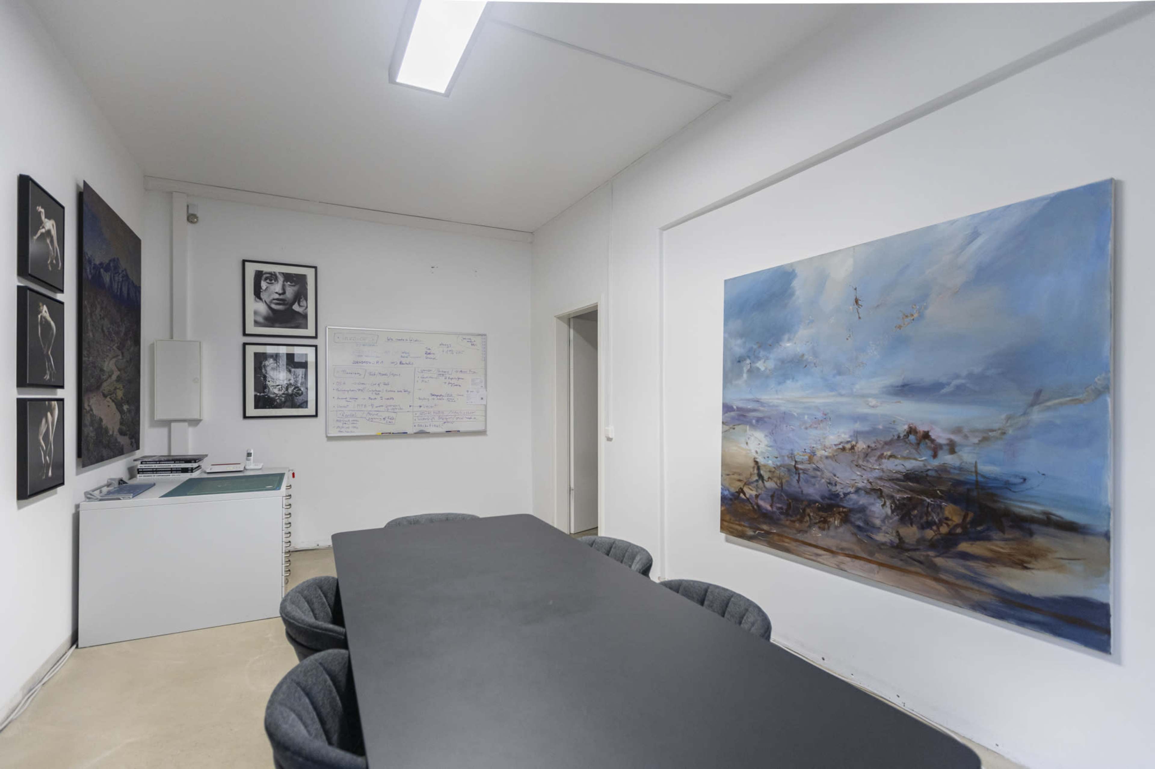 The image displays a minimalist meeting room featuring a large black table surrounded by gray chairs, with artwork and a whiteboard on the walls.