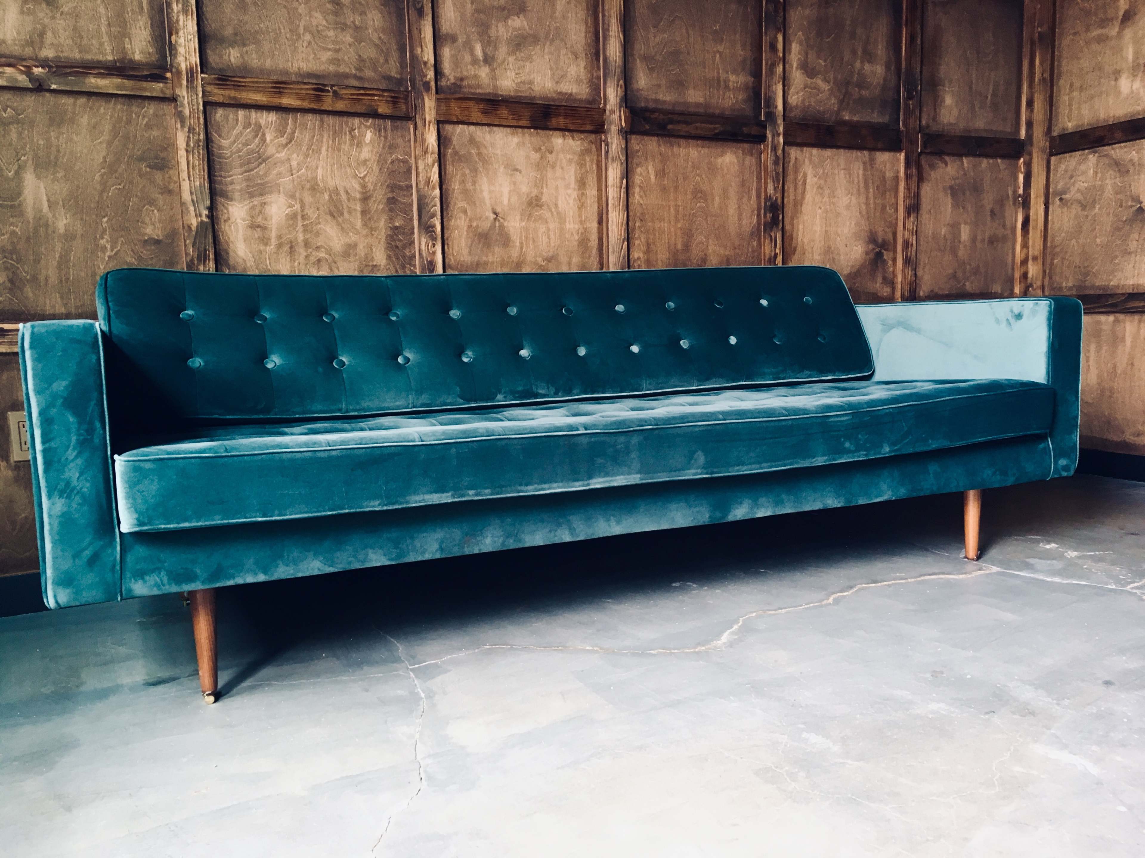 A teal velvet sofa with a button-tufted backrest and wooden legs sits against a rustic wooden wall.