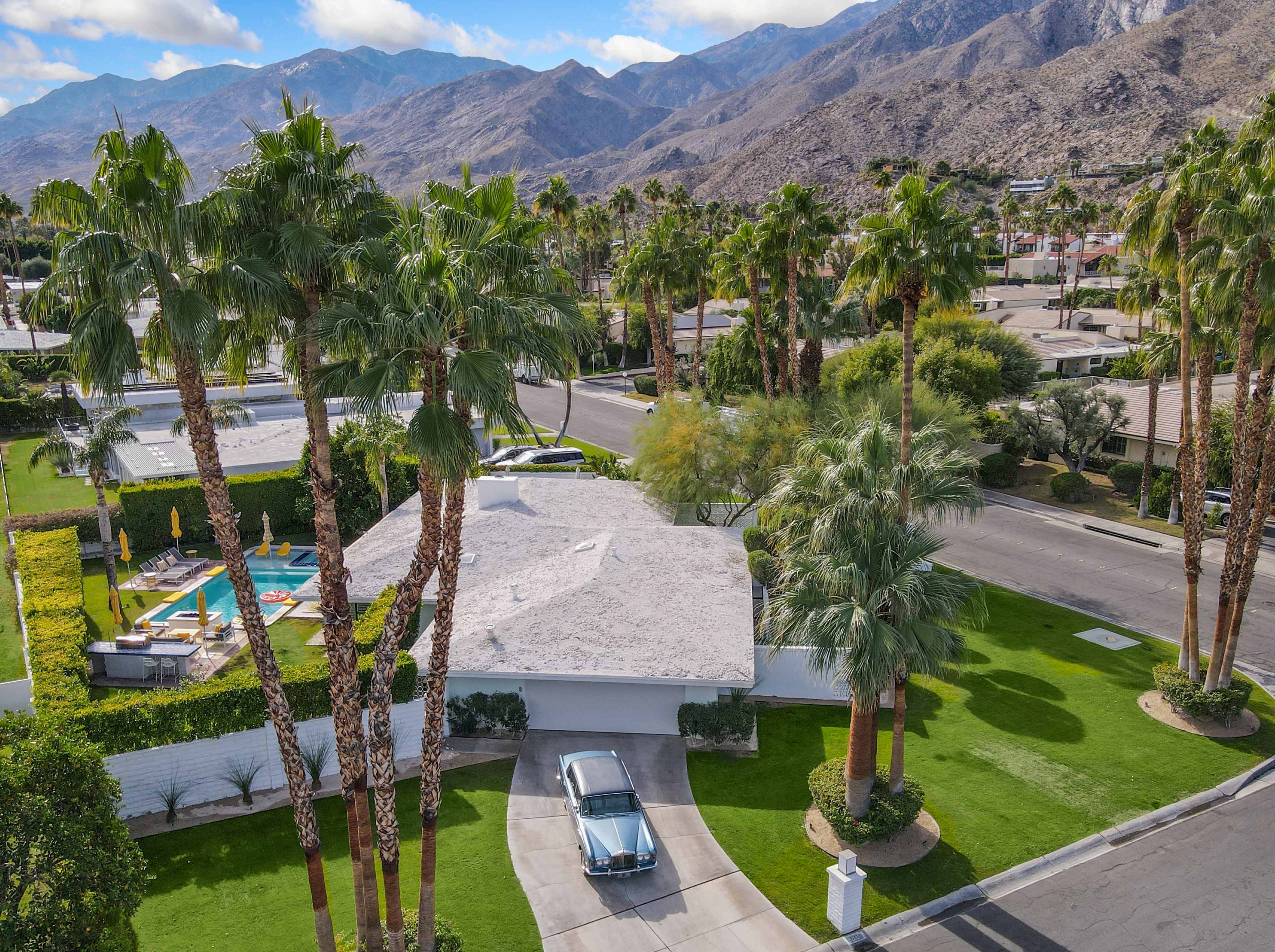 A vintage car is parked in the driveway of a modern house surrounded by palm trees and a clear view of the mountains.