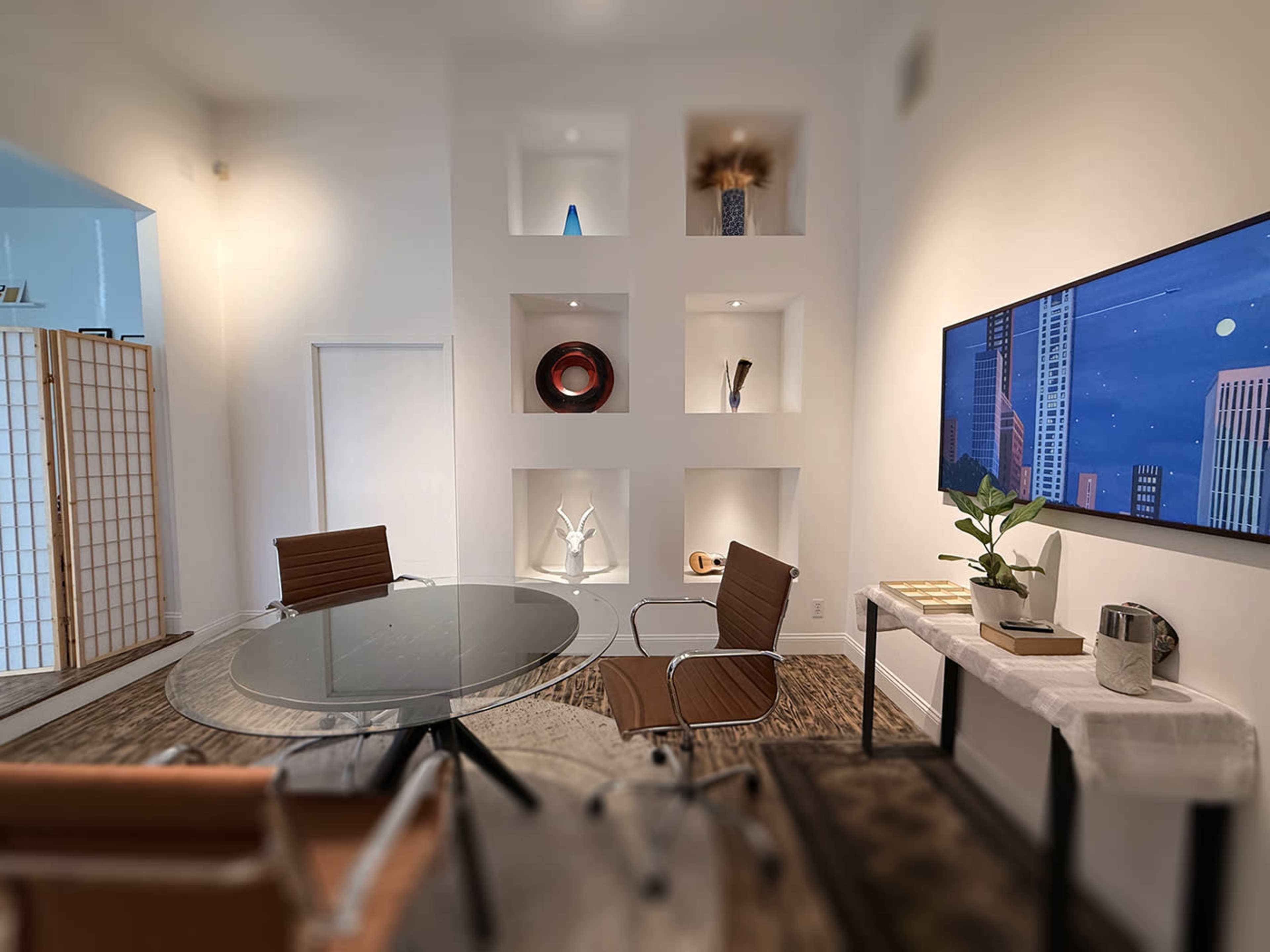 The image shows a modern conference room with a glass table and leather chairs, complemented by minimalistic shelves featuring decorative items and a large wall-mounted TV displaying a cityscape.