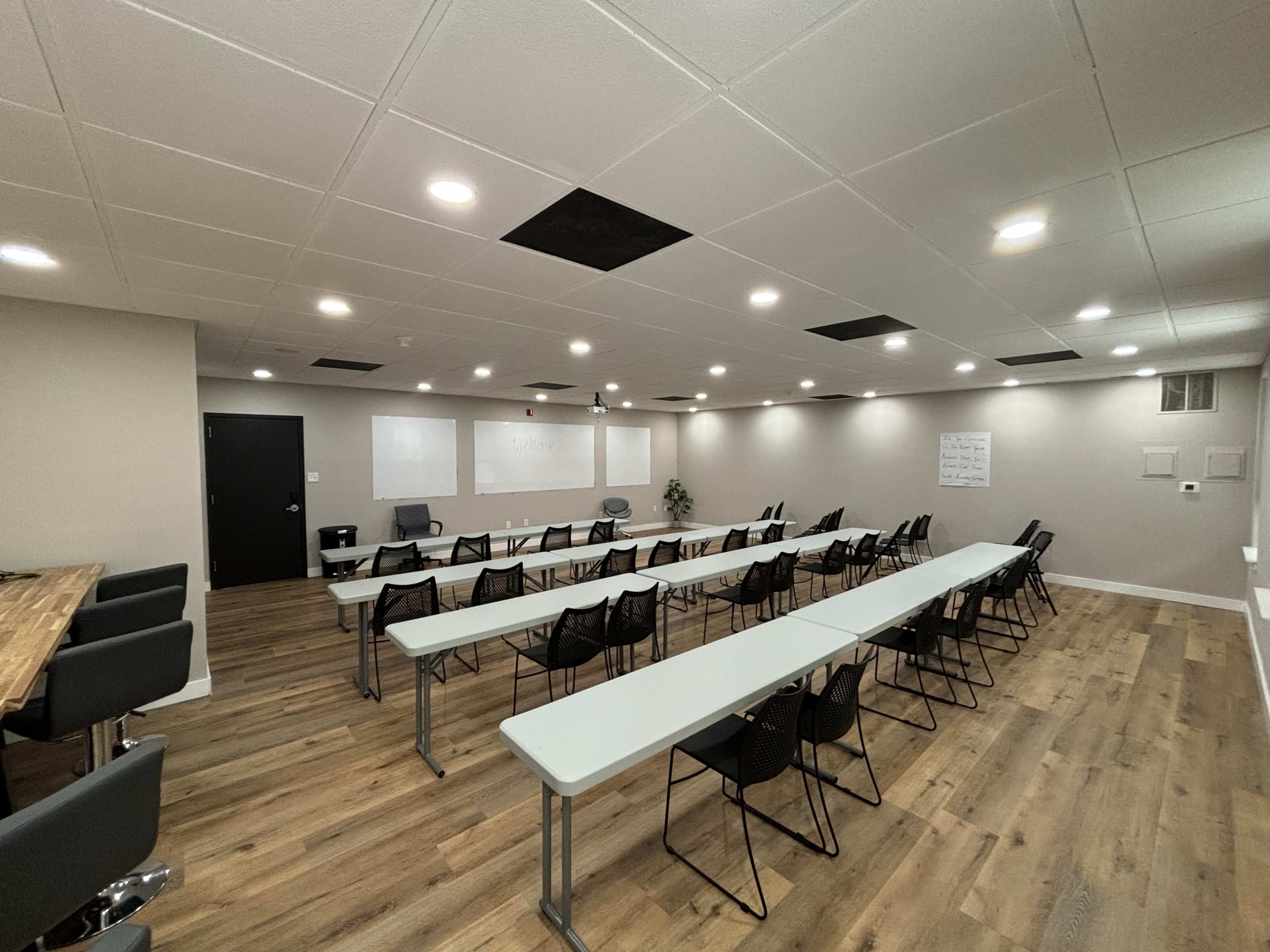 A modern conference room with long tables arranged in rows and chairs facing a blank whiteboard.