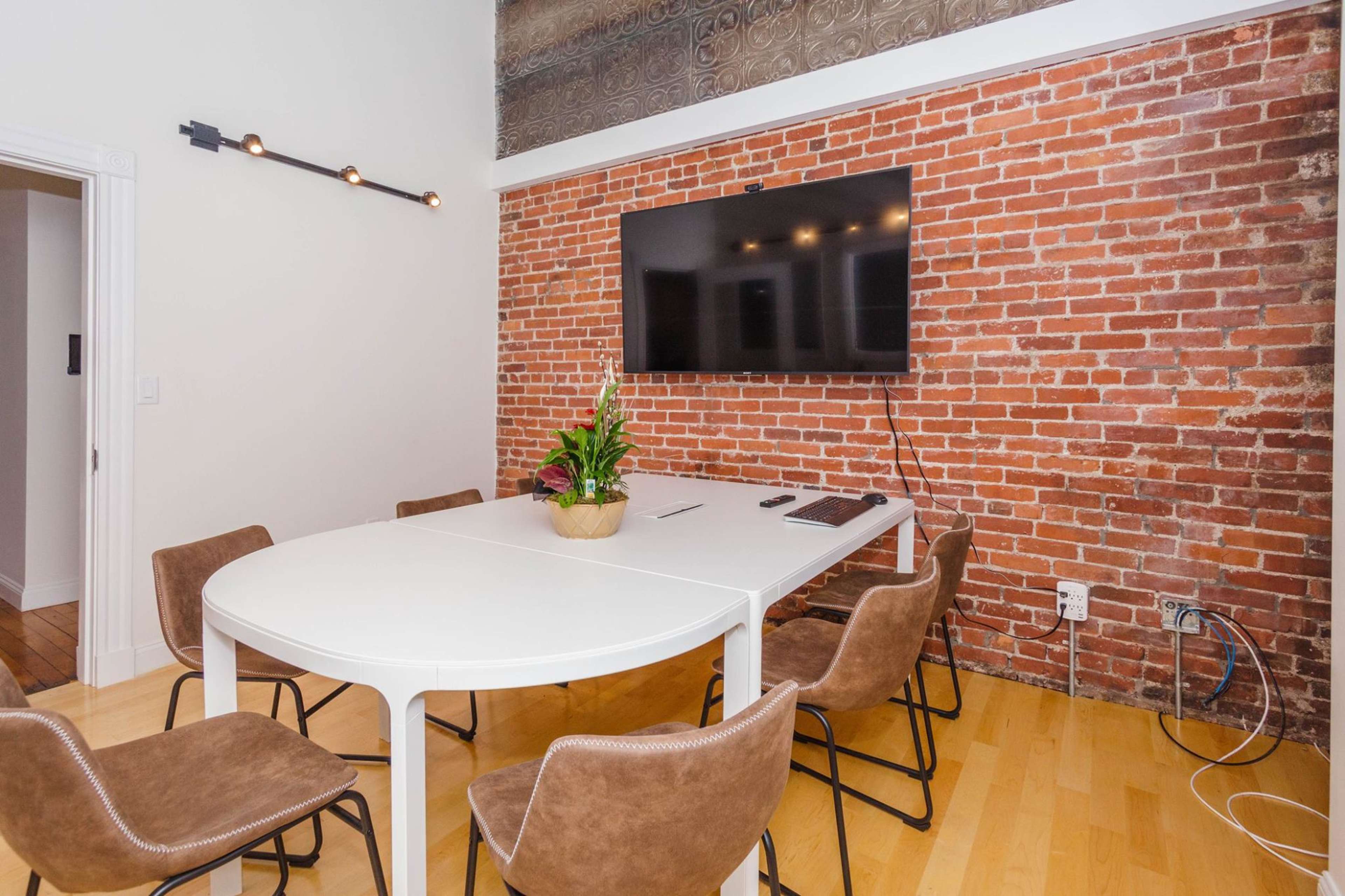A conference room with a white table, brown chairs, a plant centerpiece, and a wall-mounted television on a brick wall.