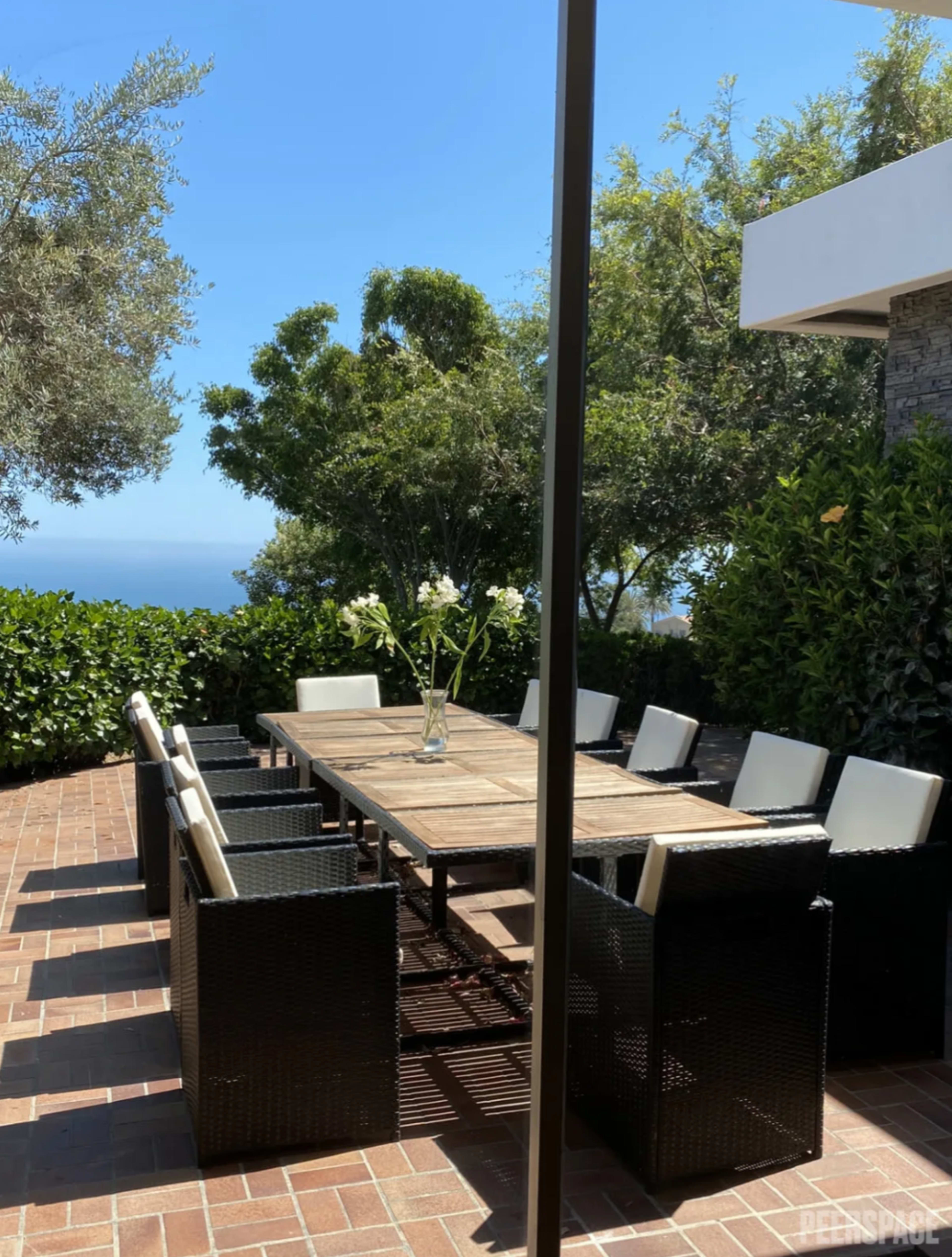 A wooden dining table surrounded by black wicker chairs sits in a landscaped outdoor area with views of greenery and the ocean.