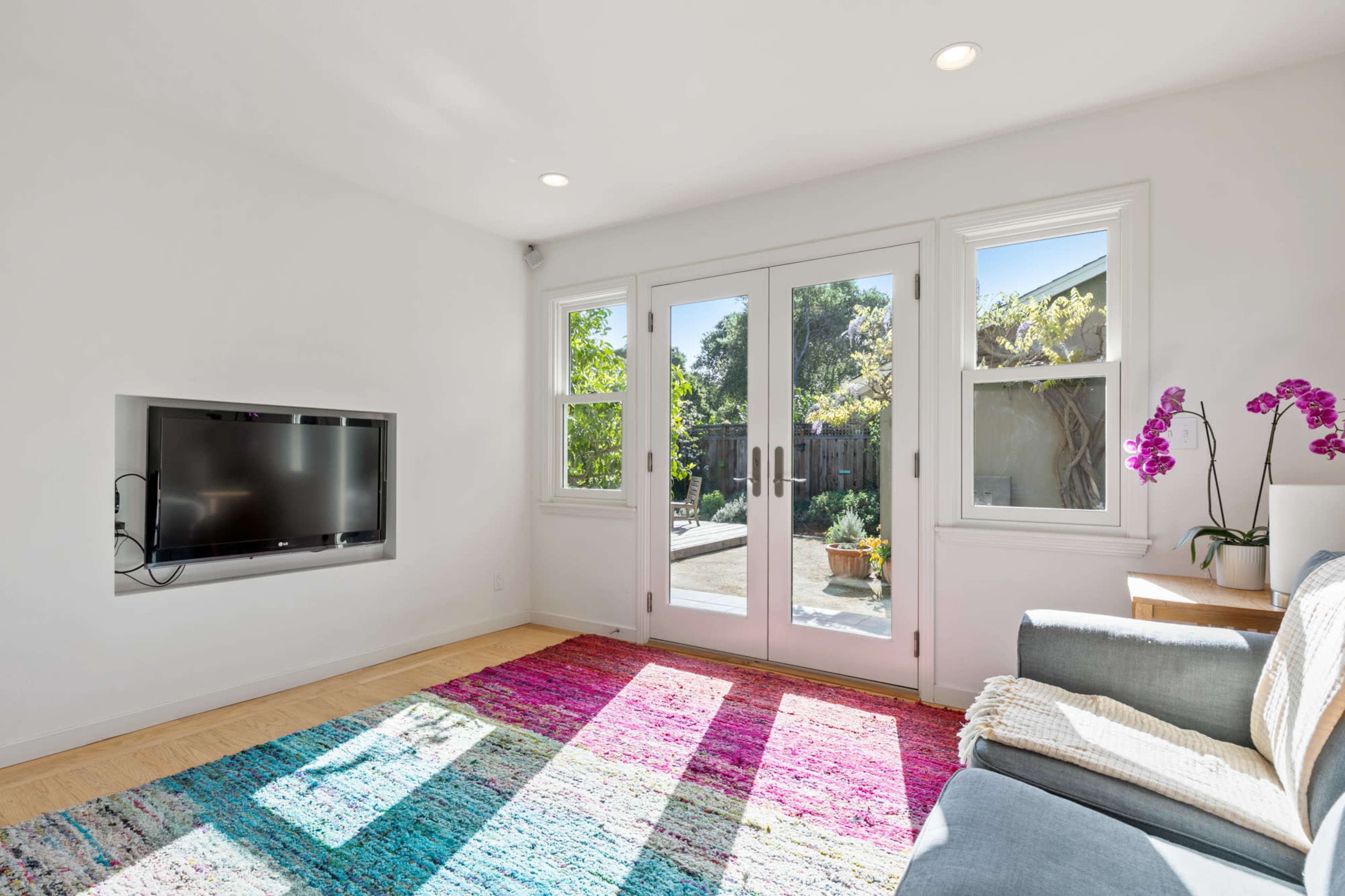 A bright living room features a mounted television, a gray sofa, and large glass doors that open to a patio surrounded by greenery.