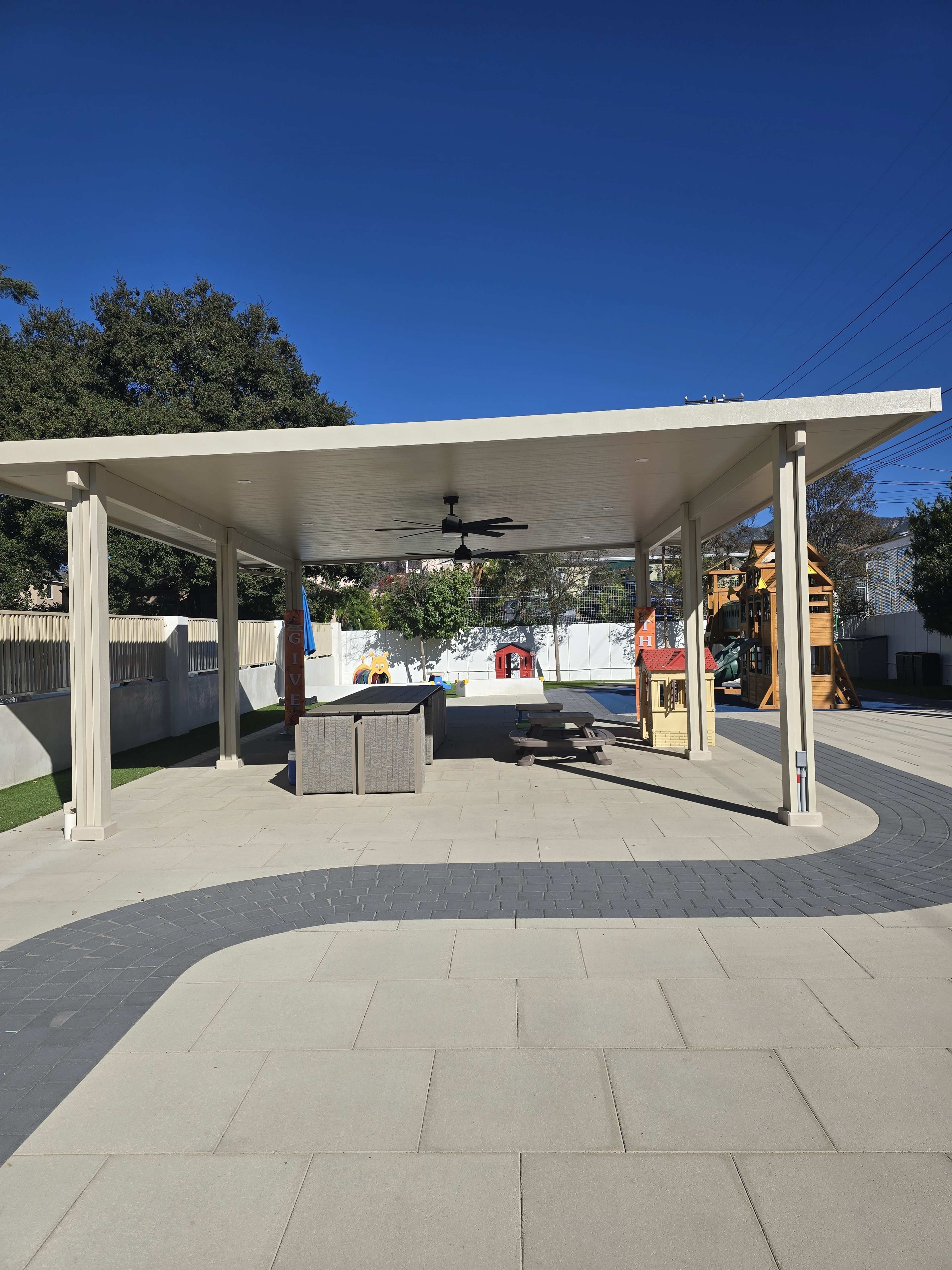 The image shows a large outdoor pavilion with a ceiling fan, overlooking a paved area and playground equipment in the background.