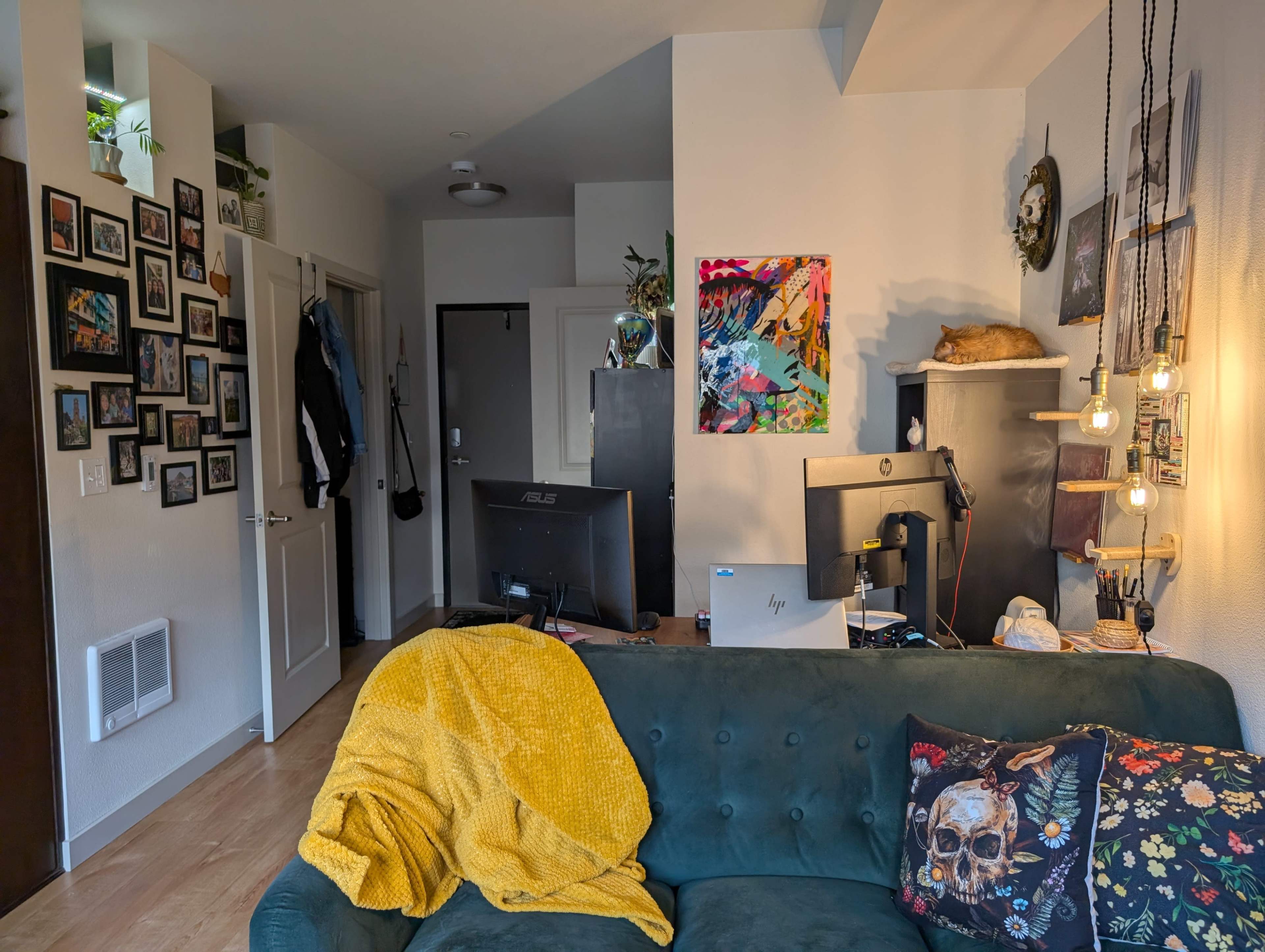 A cozy living room features a green couch with a yellow throw, surrounded by a gallery wall of framed photos and artworks, a desk with computers, and a doorway leading to another room.
