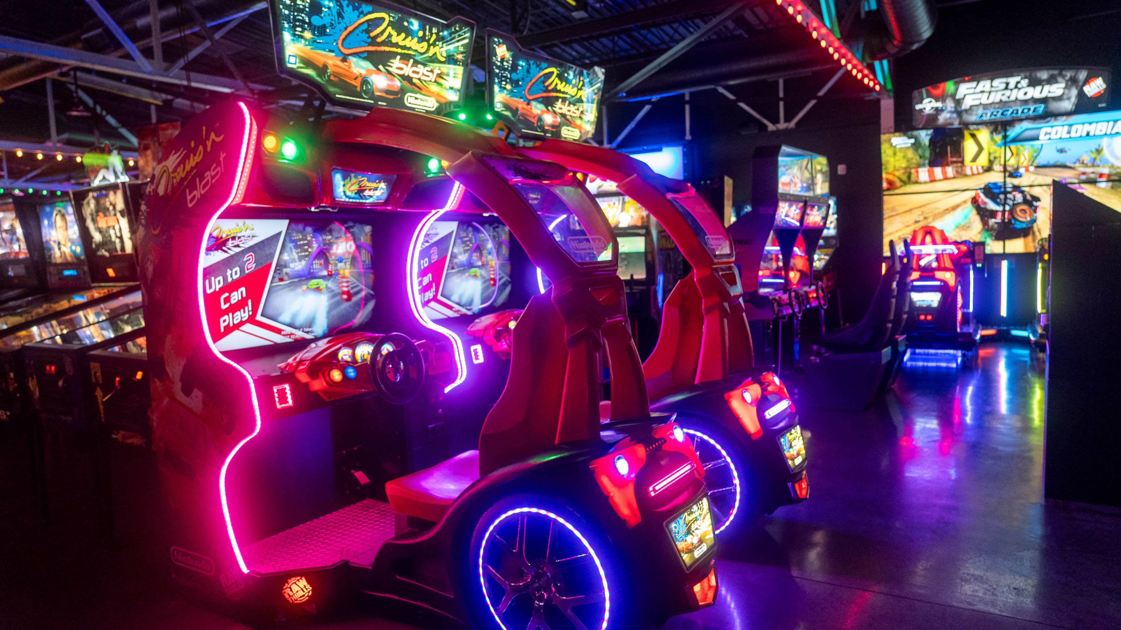 The image shows two brightly lit racing arcade game machines in an entertainment venue filled with various other arcade games.
