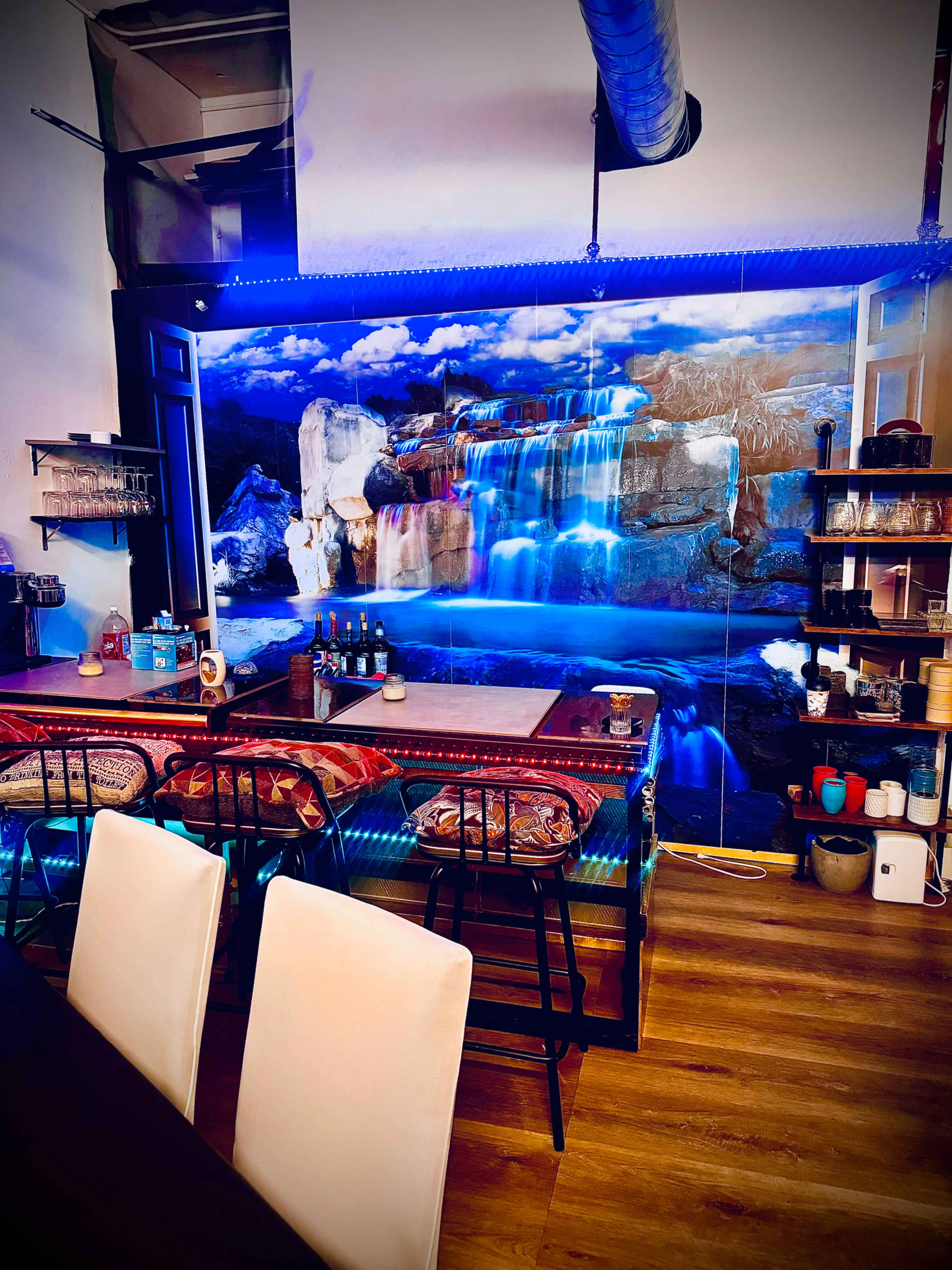The image shows a modern interior with a bar area featuring a mural of a waterfall and a counter with colorful cushions on the stools.
