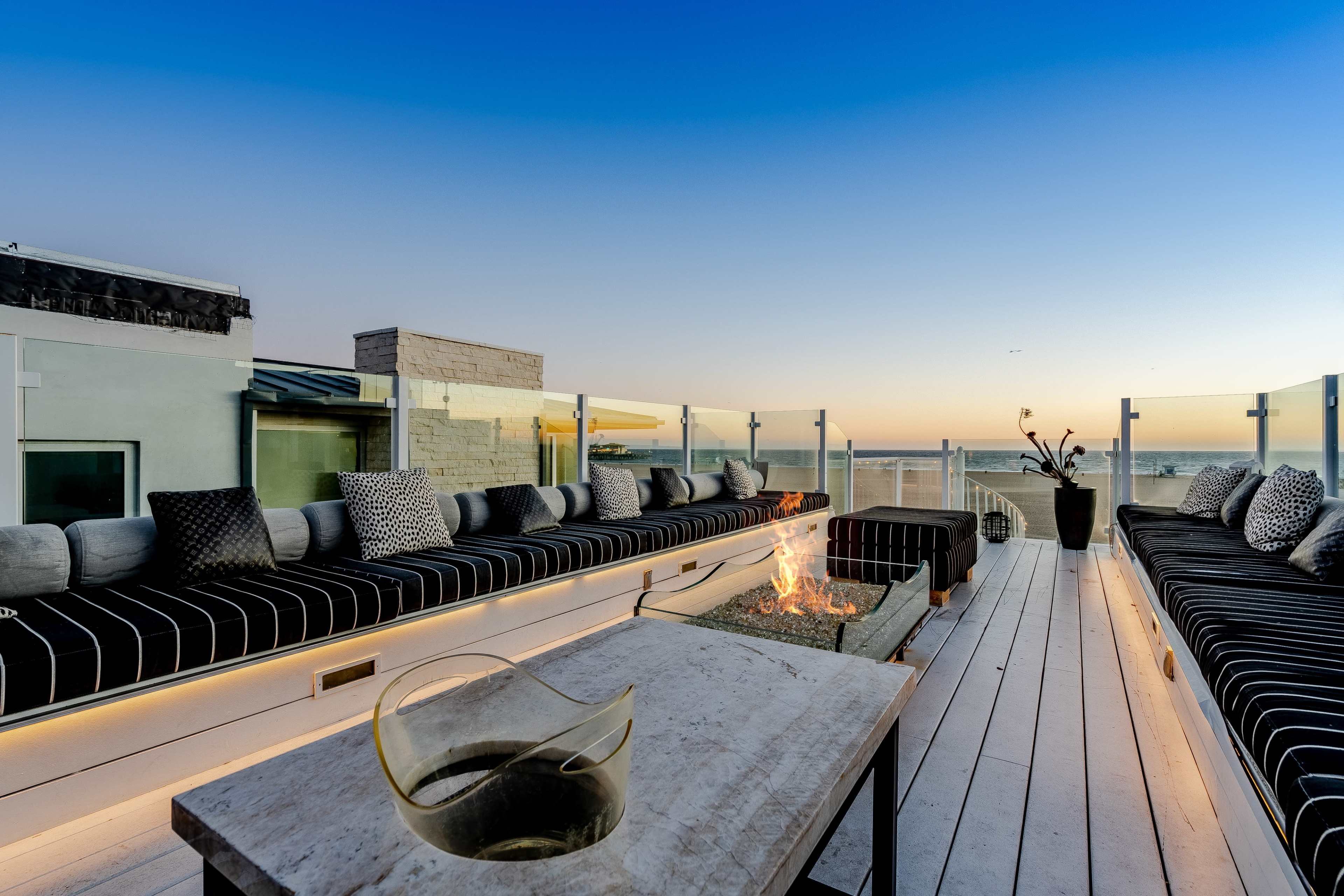 The image shows a rooftop terrace featuring a long seating area with striped cushions, a fire pit in the center, and a clear view of the horizon at sunset.