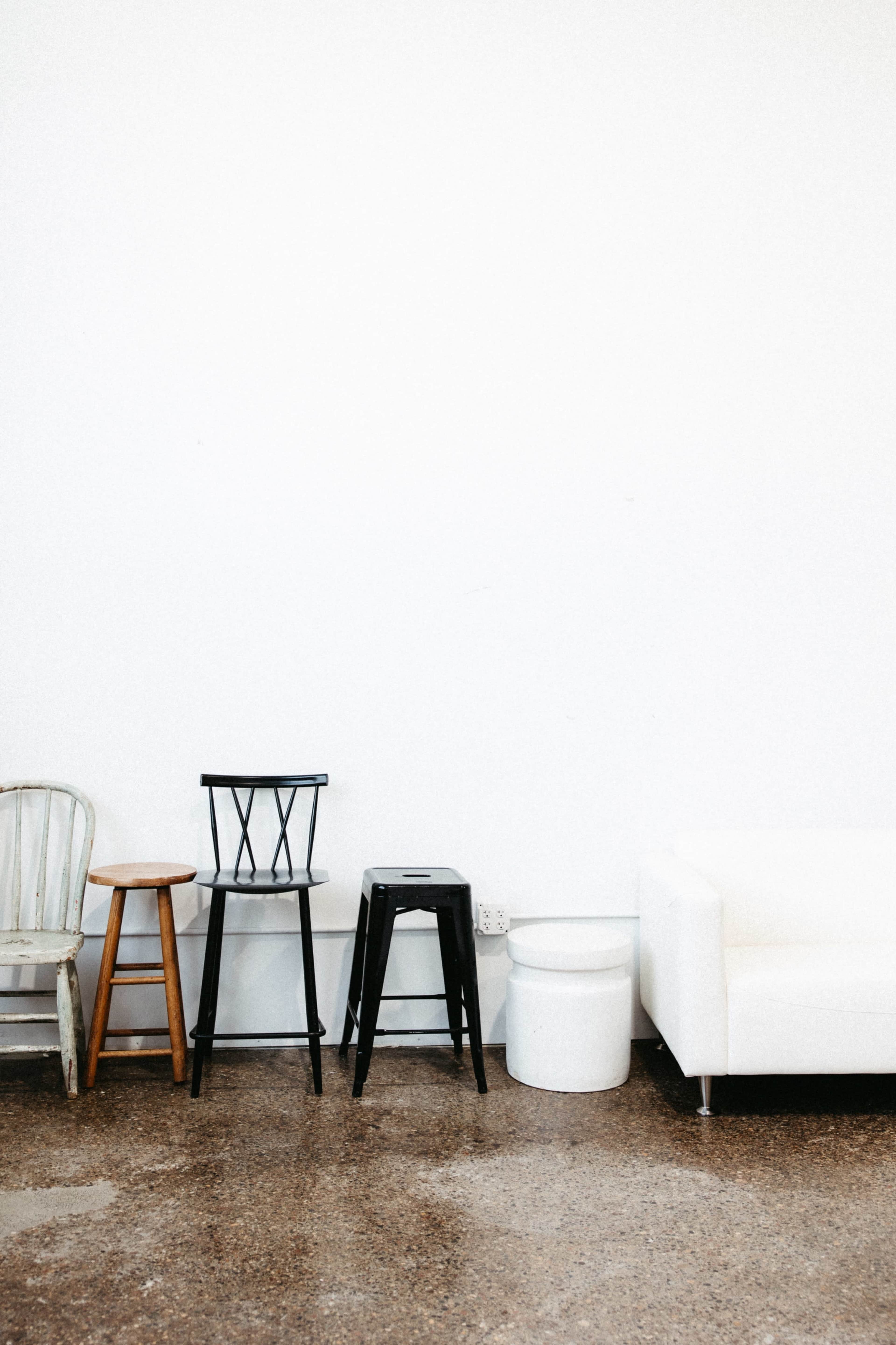 The image shows a minimalist interior with three wooden stools and a white sofa arranged against a white wall.