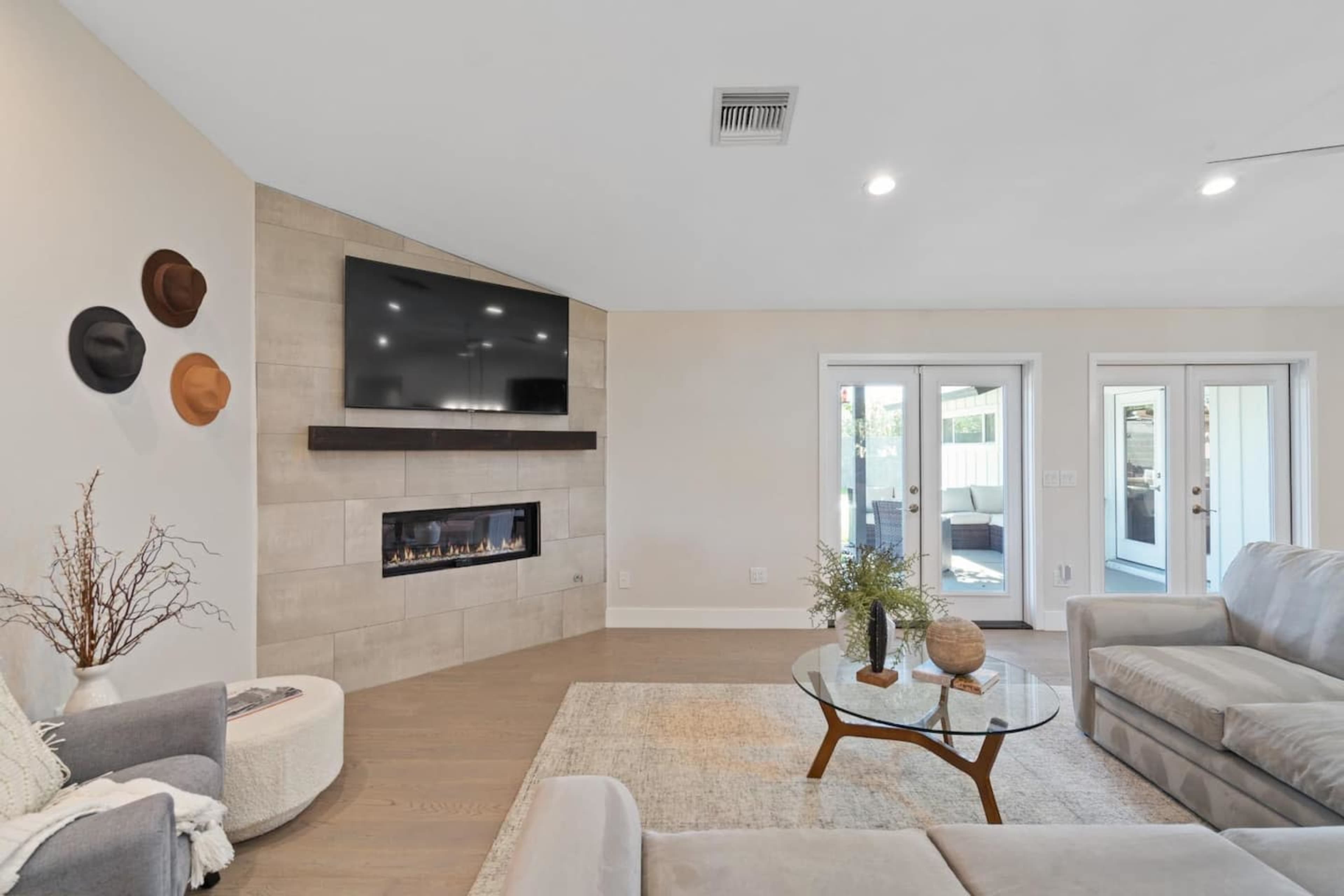 A modern living room features a sleek wall-mounted TV above a linear fireplace, two sofas, a glass coffee table, and large sliding doors leading to an outdoor space.