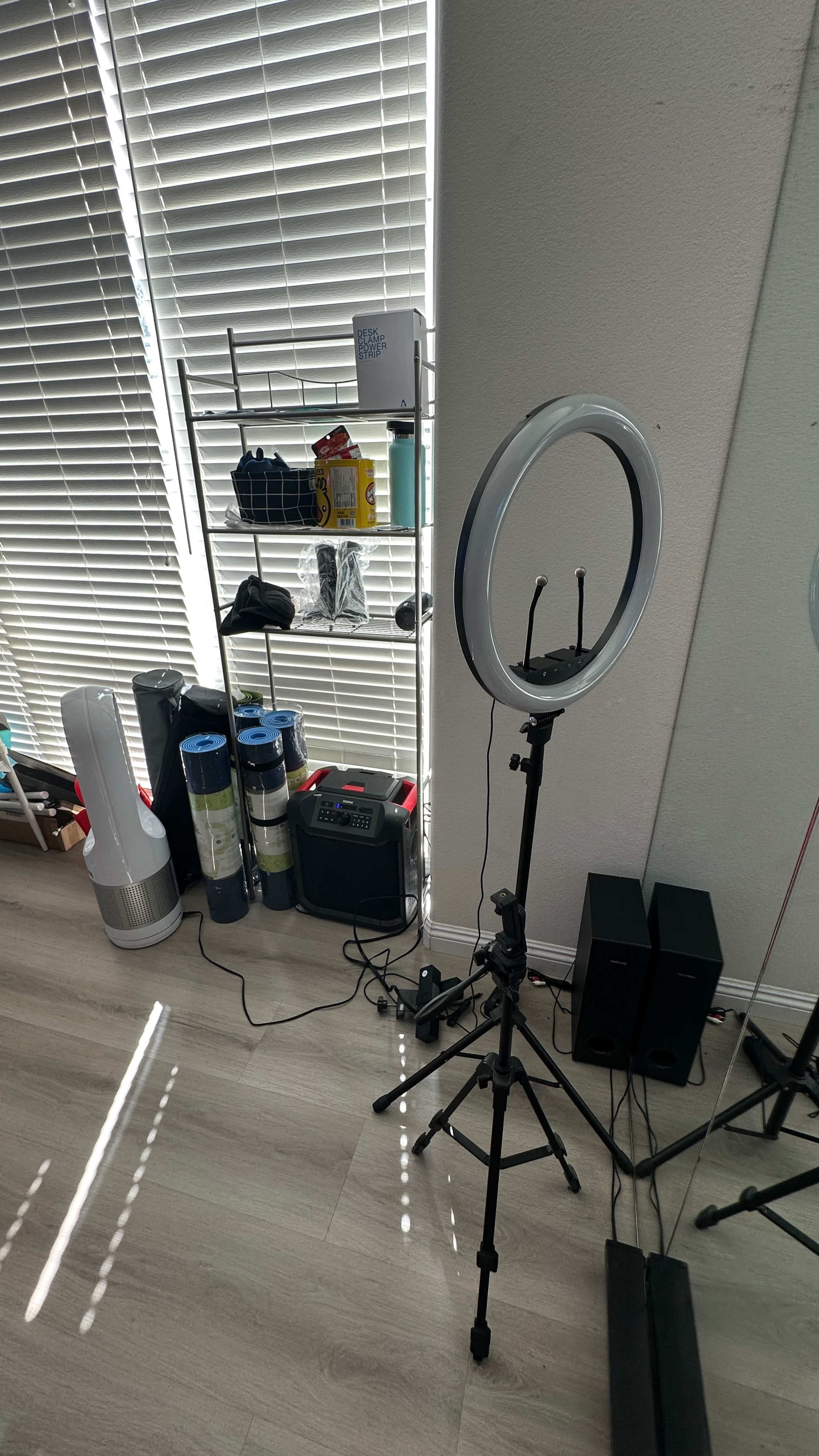 A ring light on a stand positioned near a shelf containing various items, with a mirror reflecting the scene and sunlight filtering through window blinds.