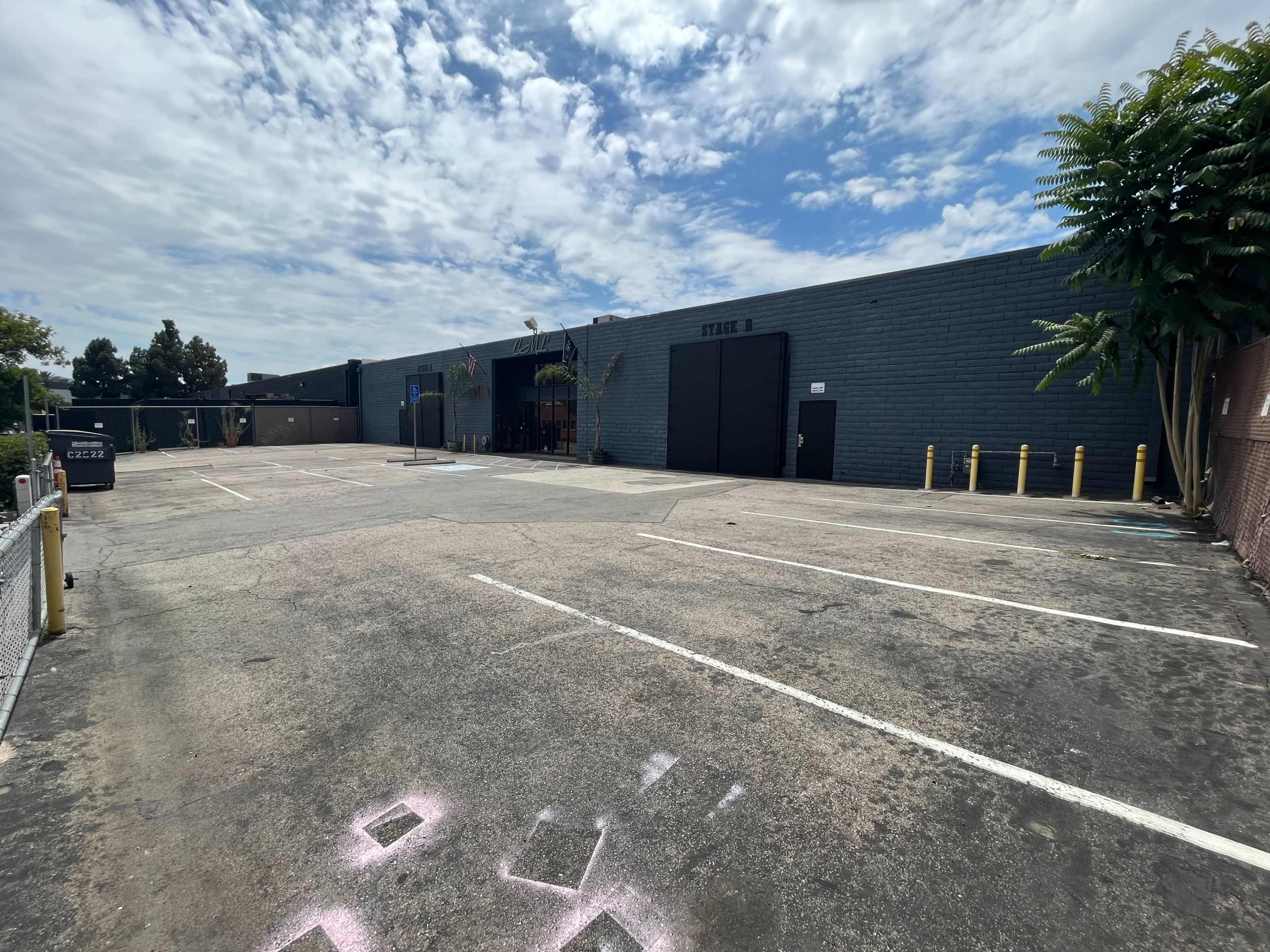 The image shows a large, empty parking lot in front of a gray building under a partially cloudy sky.