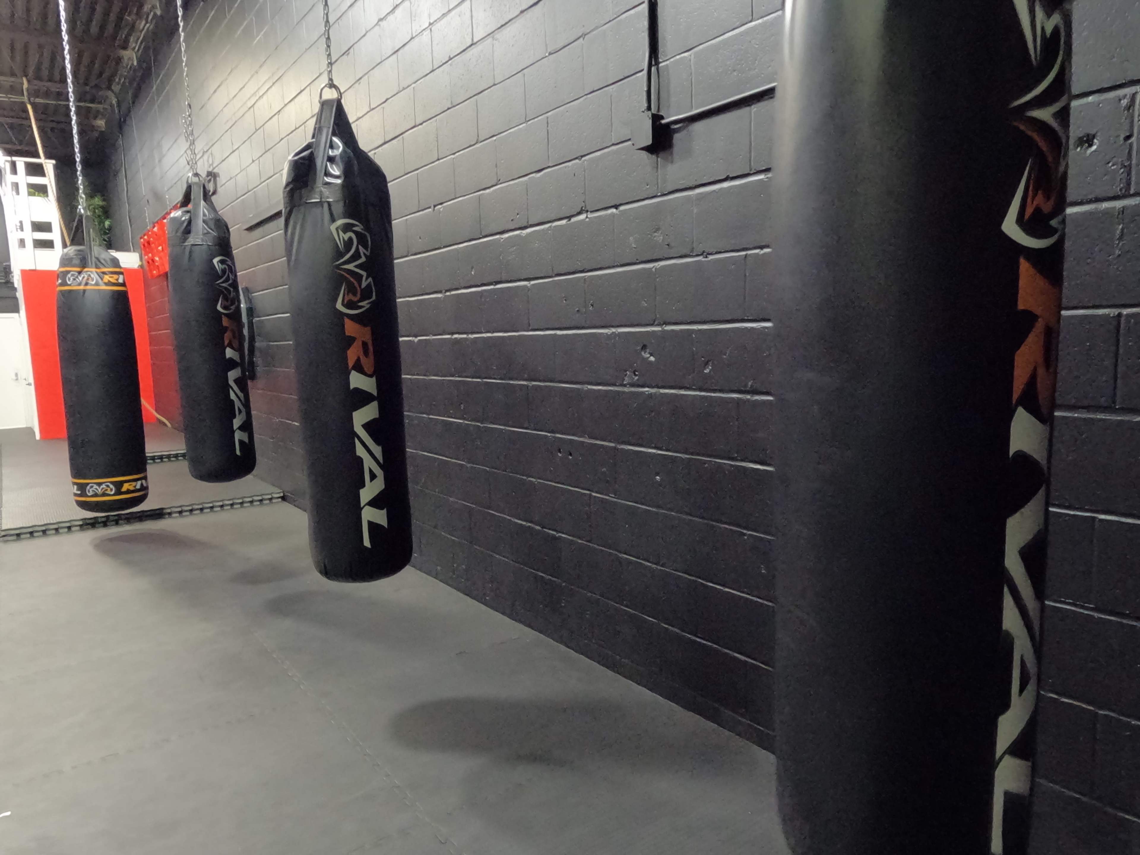 Three hanging boxing bags are suspended from the ceiling in a sparsely decorated gym with black walls.