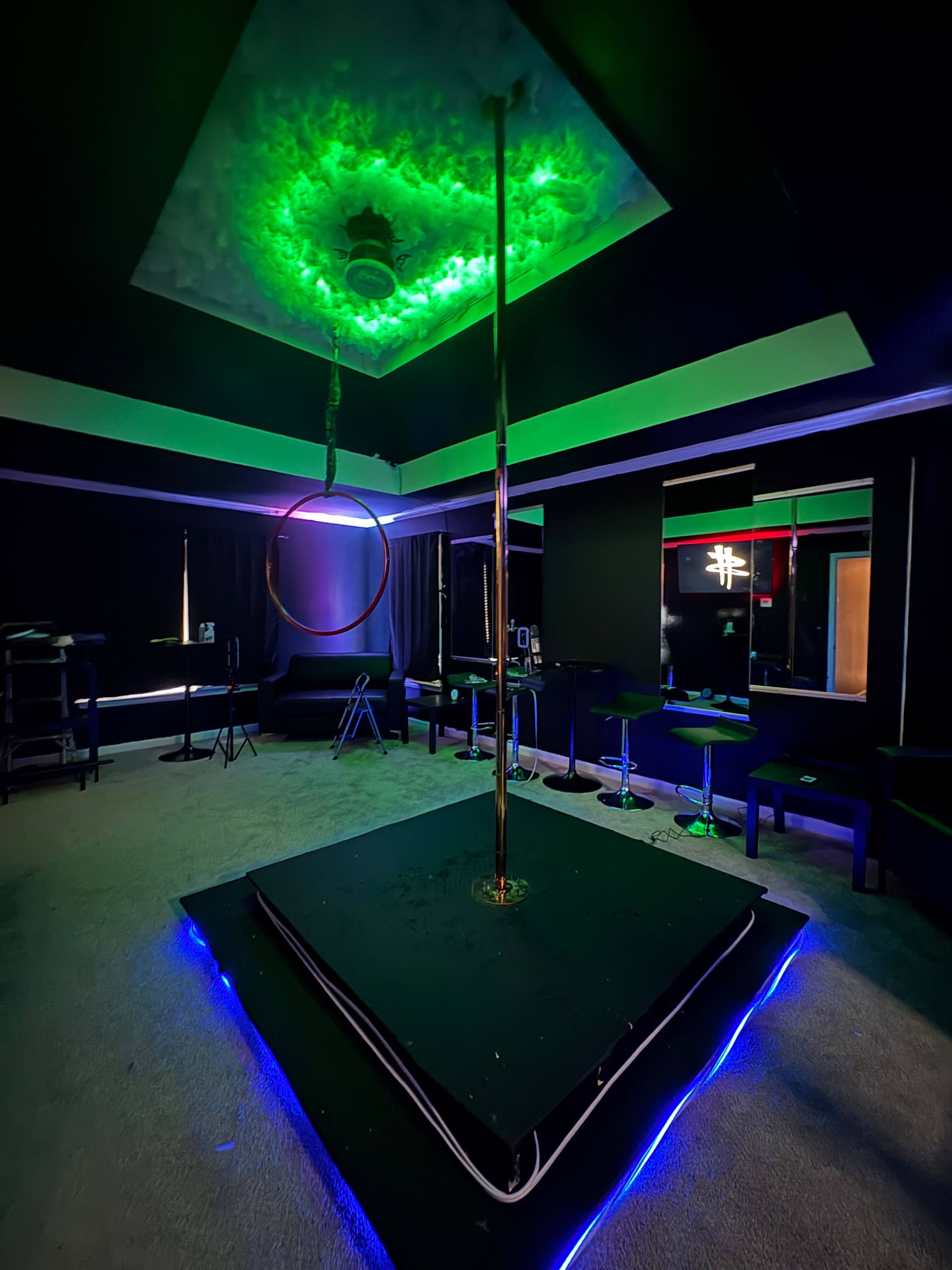 The image shows a dimly lit room featuring a circular performance stage with a pole and ring, surrounded by bar stools and illuminated by green and blue lighting.