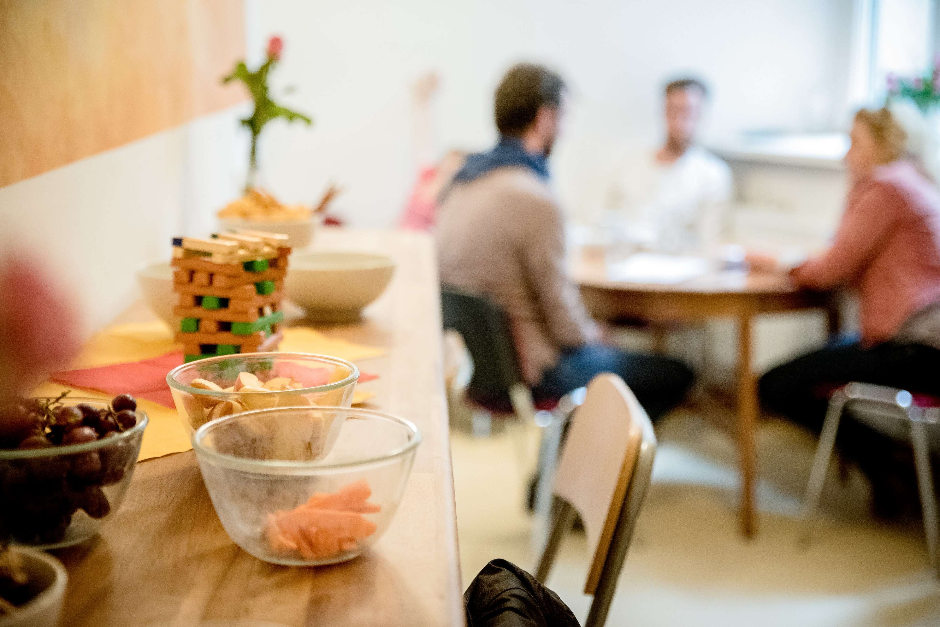 A blurred conversation takes place at a table in a kitchen, with snacks in clear bowls on a countertop in the foreground.