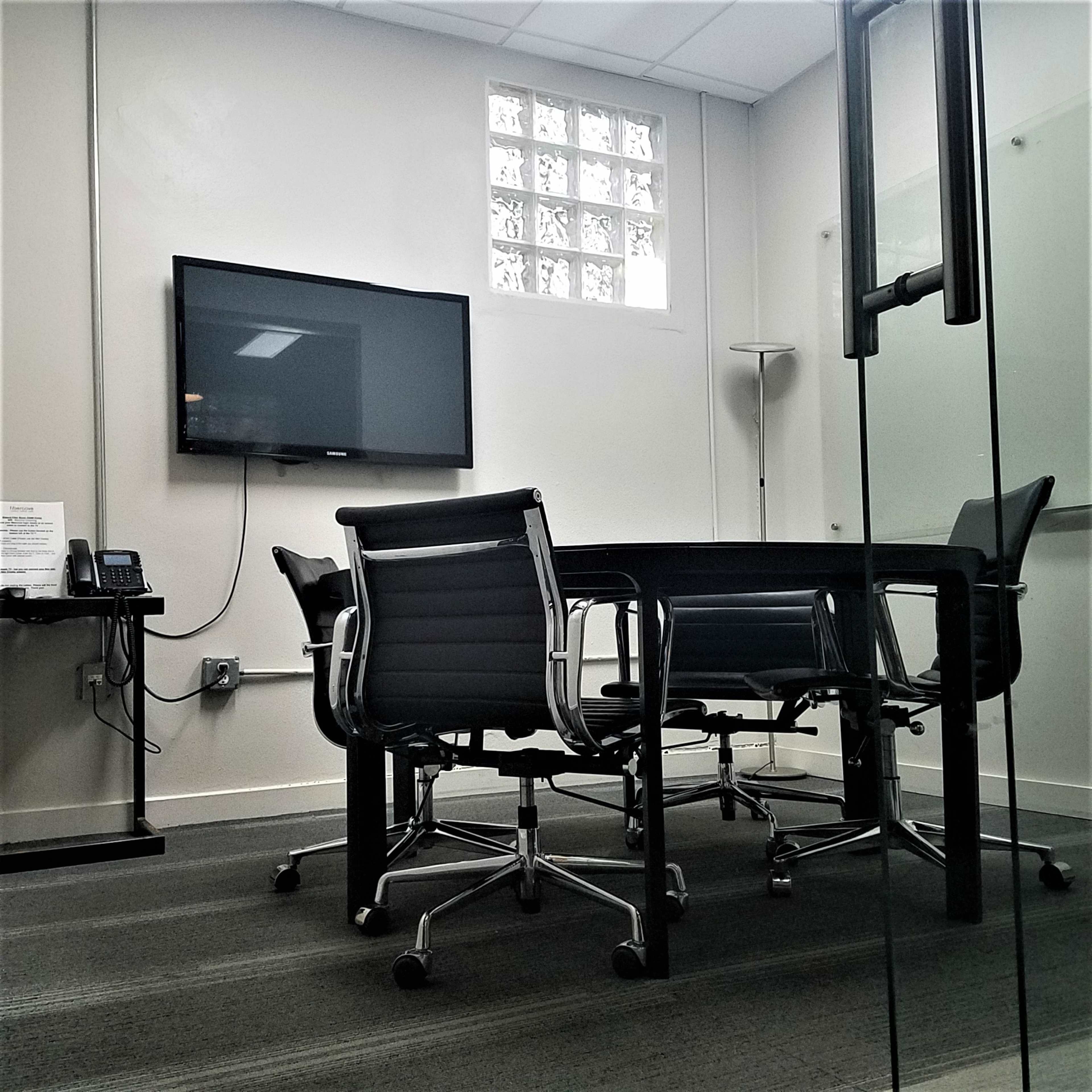 The image shows a small conference room with a round table and several black ergonomic chairs, a television mounted on the wall, and a glass partition.