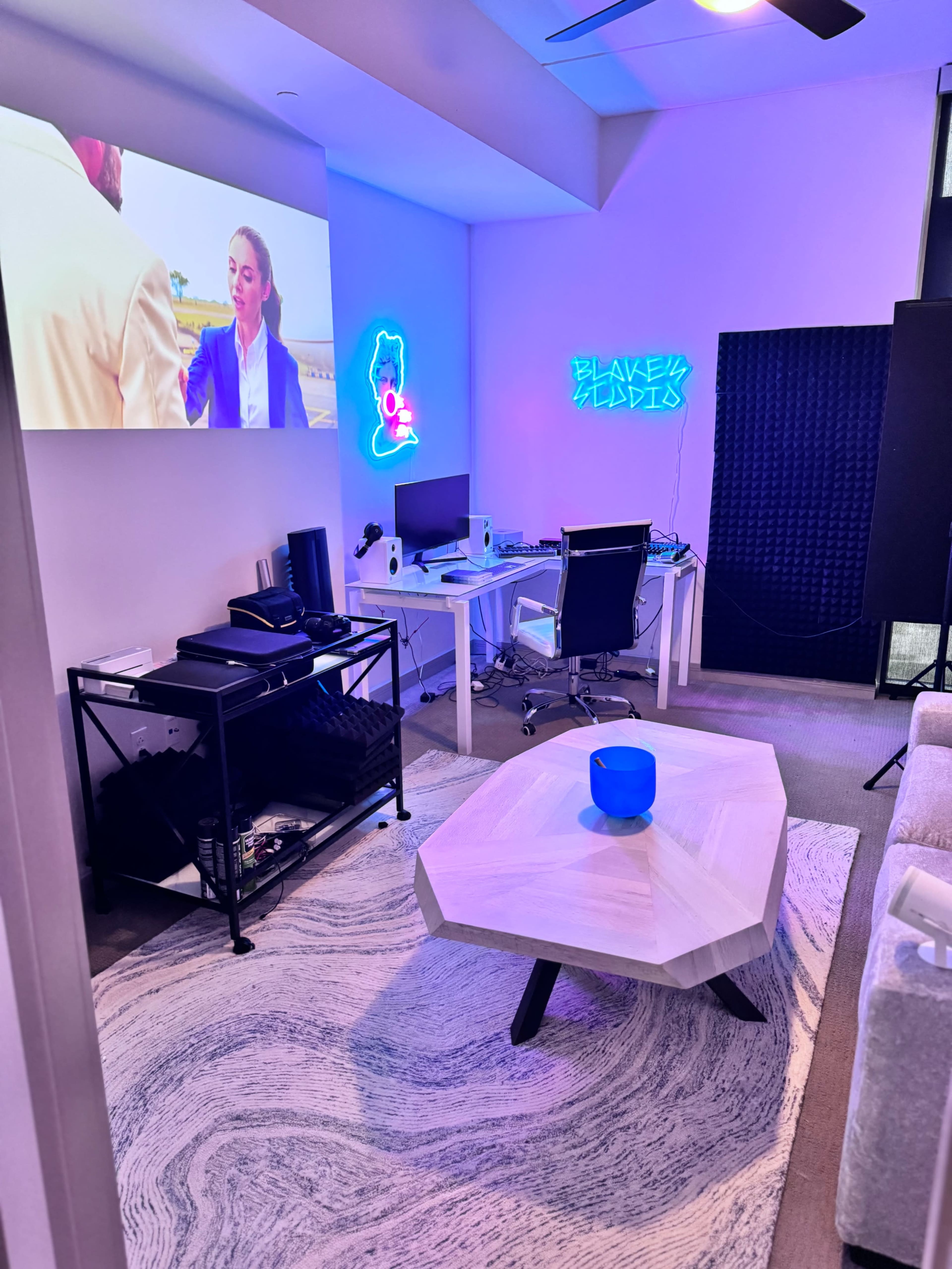 A modern workspace features a wall-mounted projection screen, two desks with computers, and a neon sign, all complemented by a stylish octagonal coffee table and a light-colored rug.