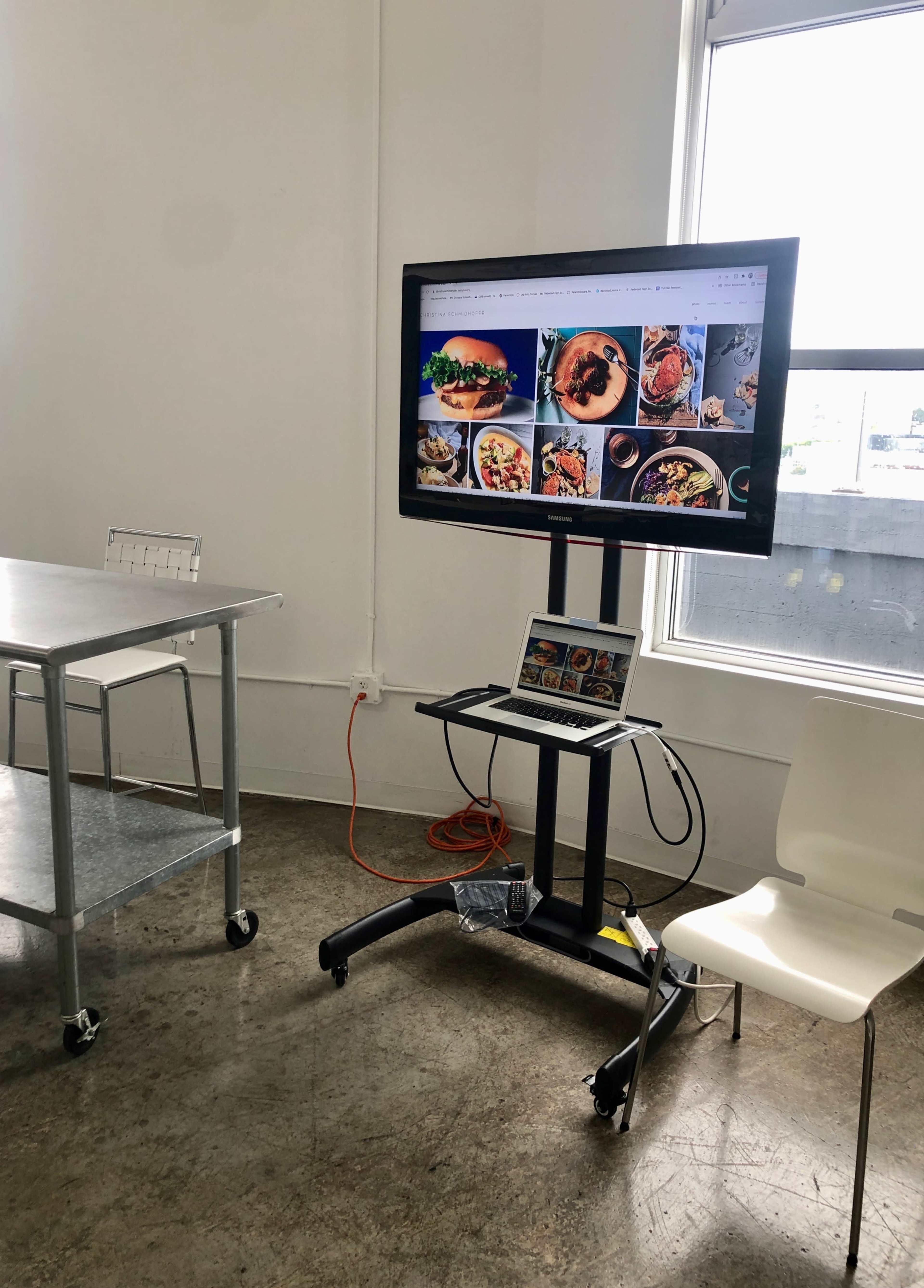 A monitor displaying food images is set up on a stand next to a metal table and a white chair in a well-lit room.