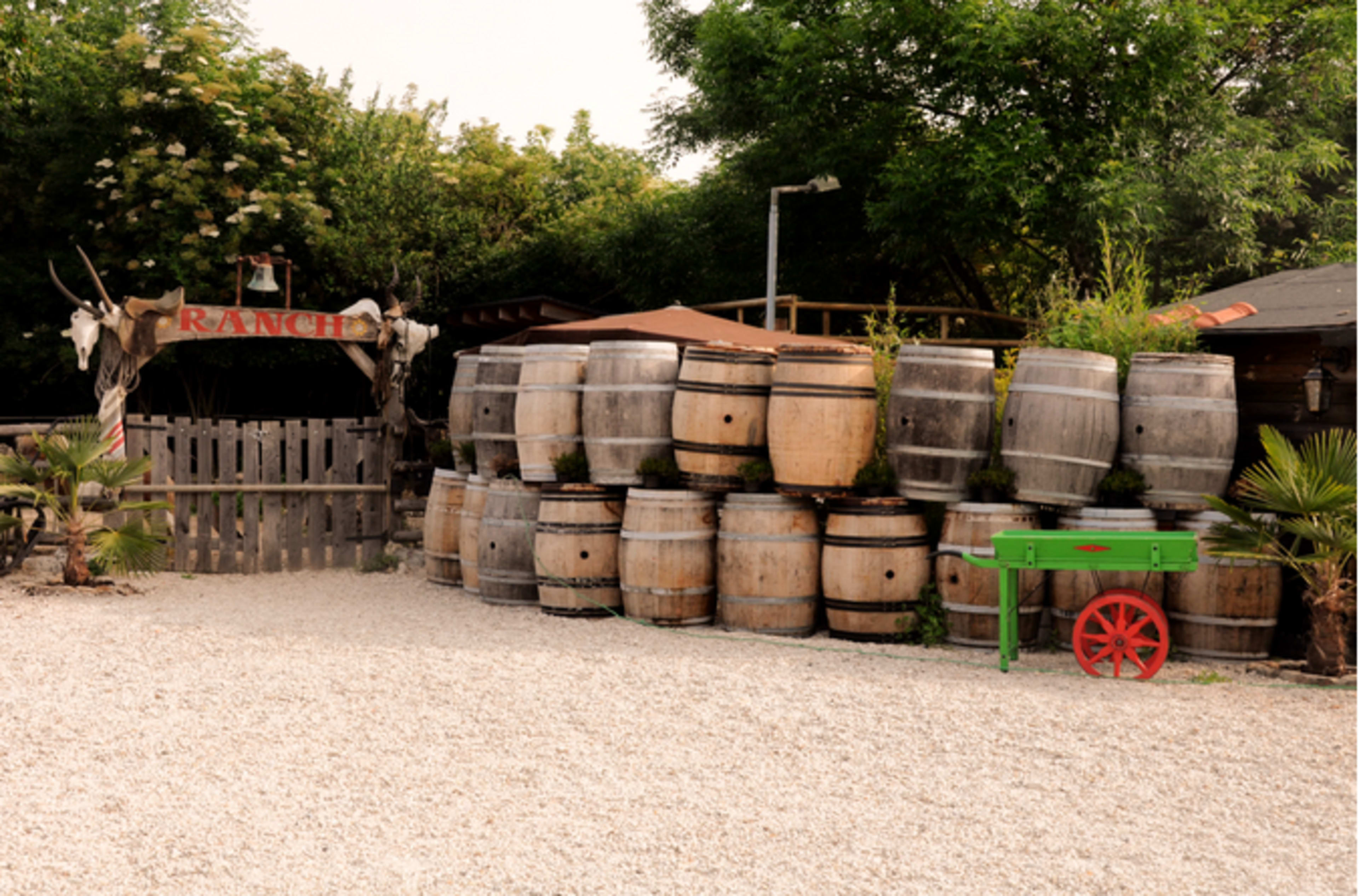 A stack of wooden barrels is positioned next to a green cart in front of a ranch entrance surrounded by trees.