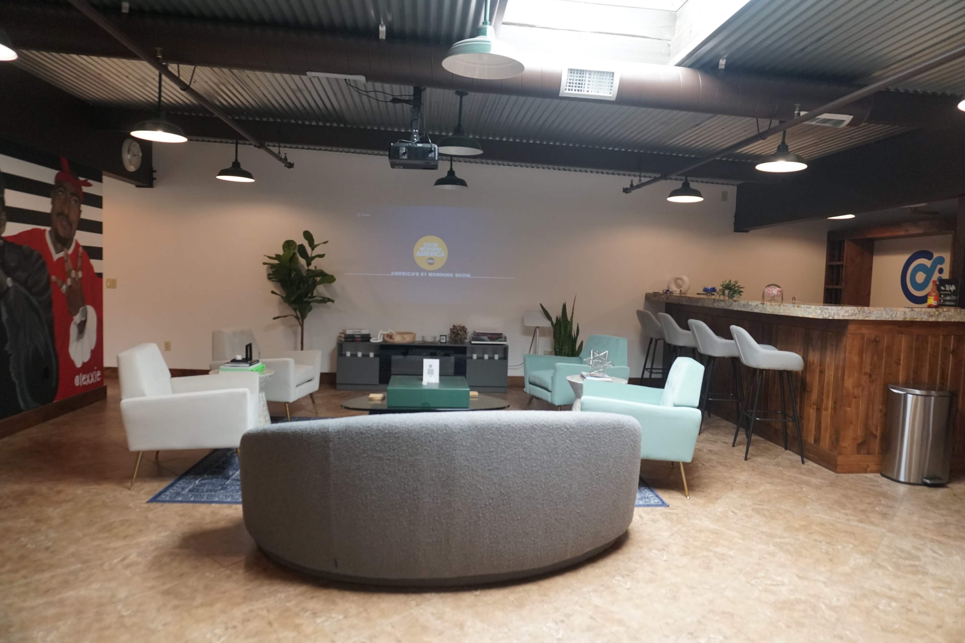 A modern lounge area with two light blue chairs, a grey sofa, a bar counter, and seating around a central coffee table under industrial-style lighting.
