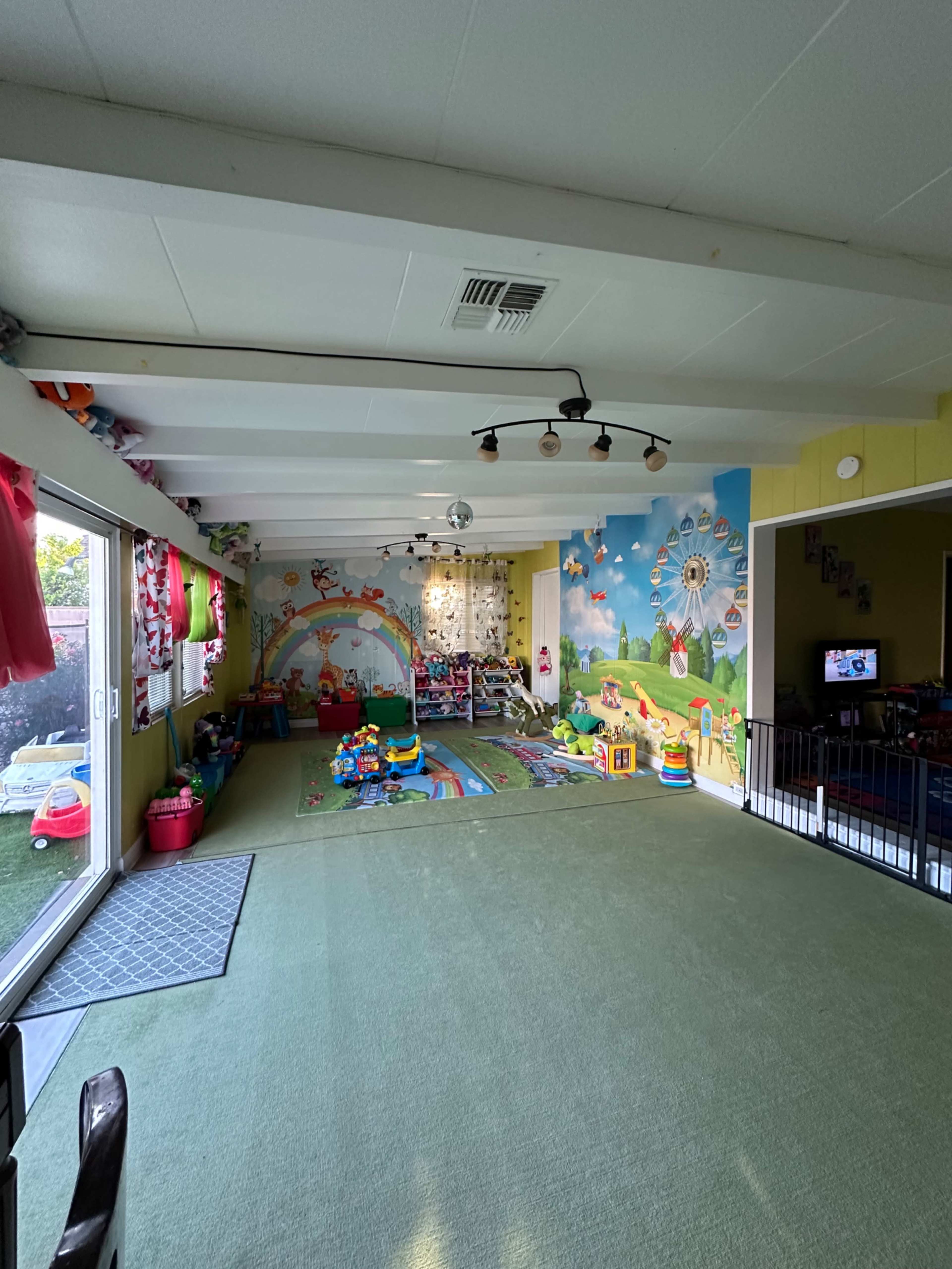 The image shows a bright, spacious playroom with colorful wall murals, play mats, toys scattered around, and sliding glass doors leading outside.