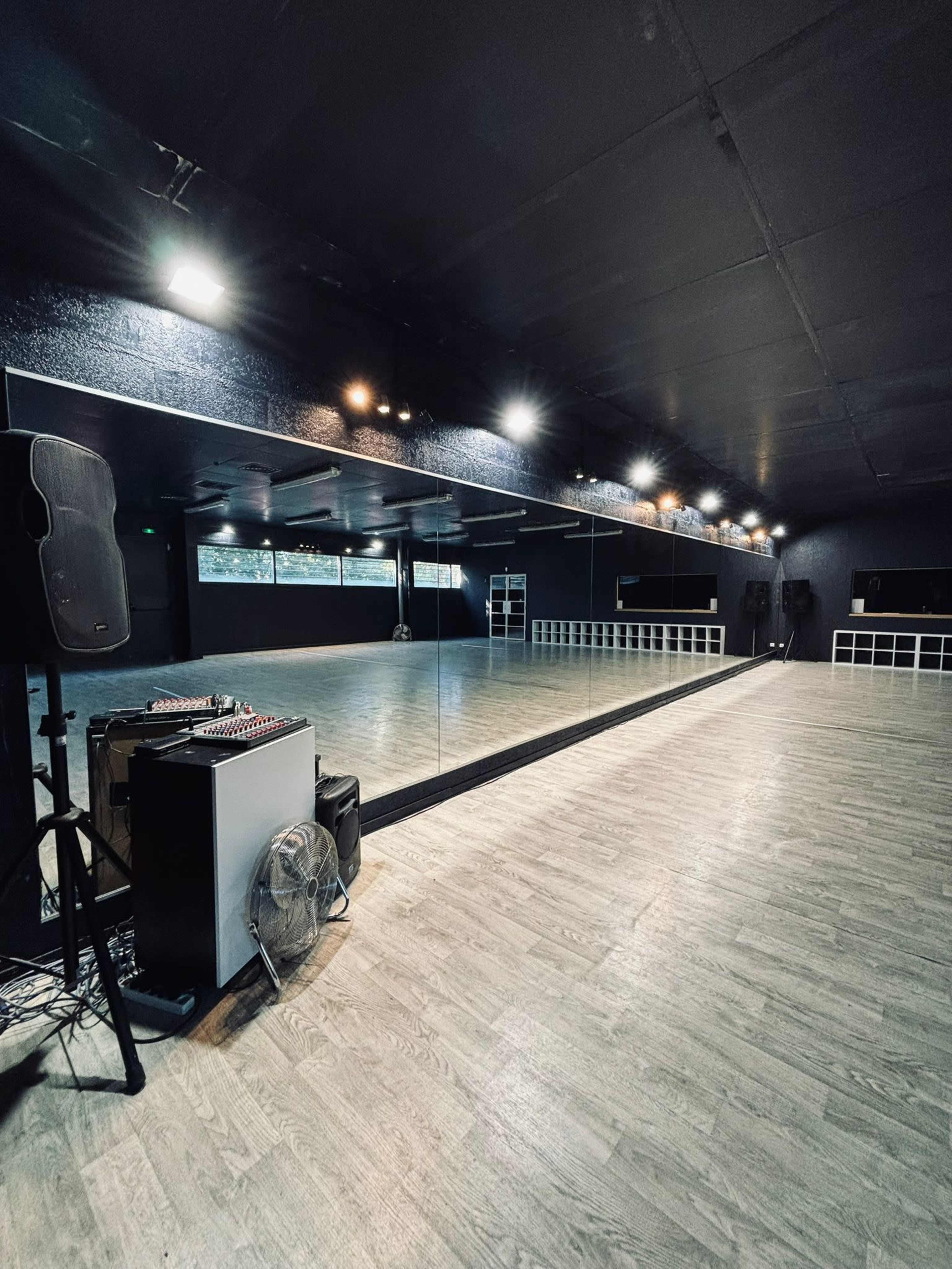 The image shows an empty dance studio featuring a large mirror, wooden floors, and sound equipment on one side.