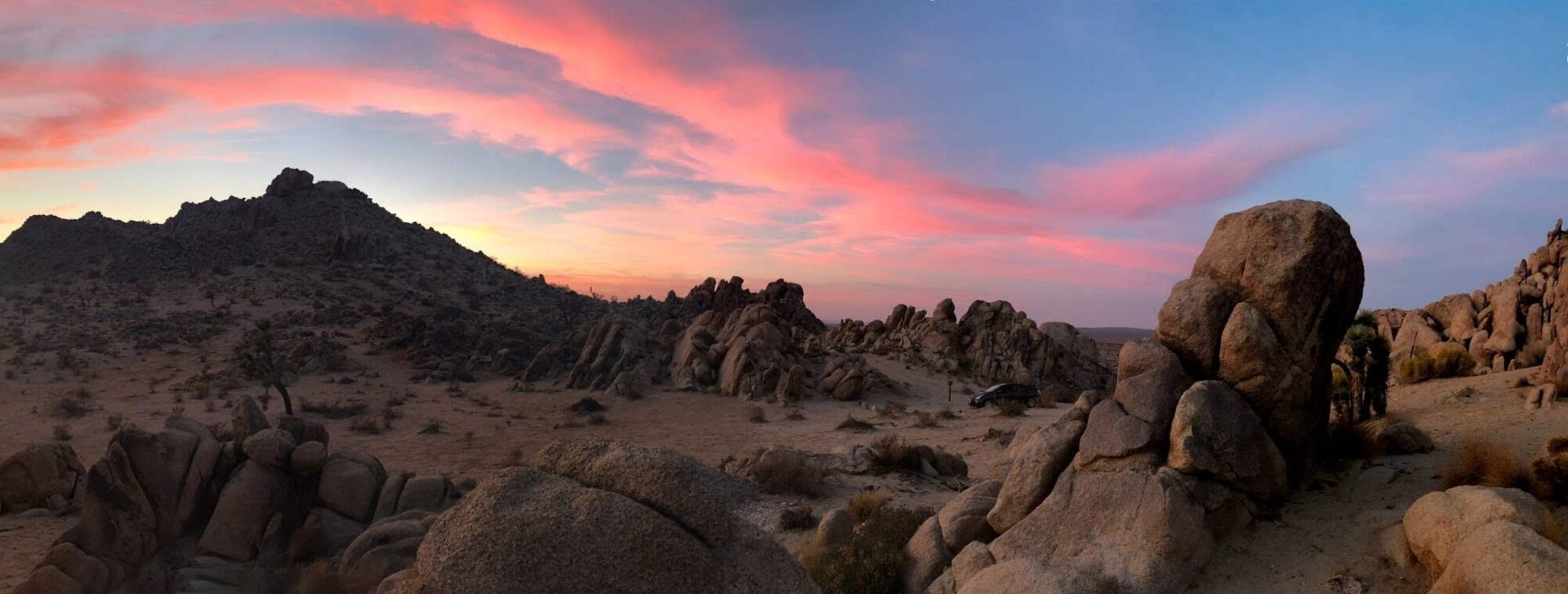The image shows a panoramic view of a desert landscape with large rock formations and a colorful sunset sky.