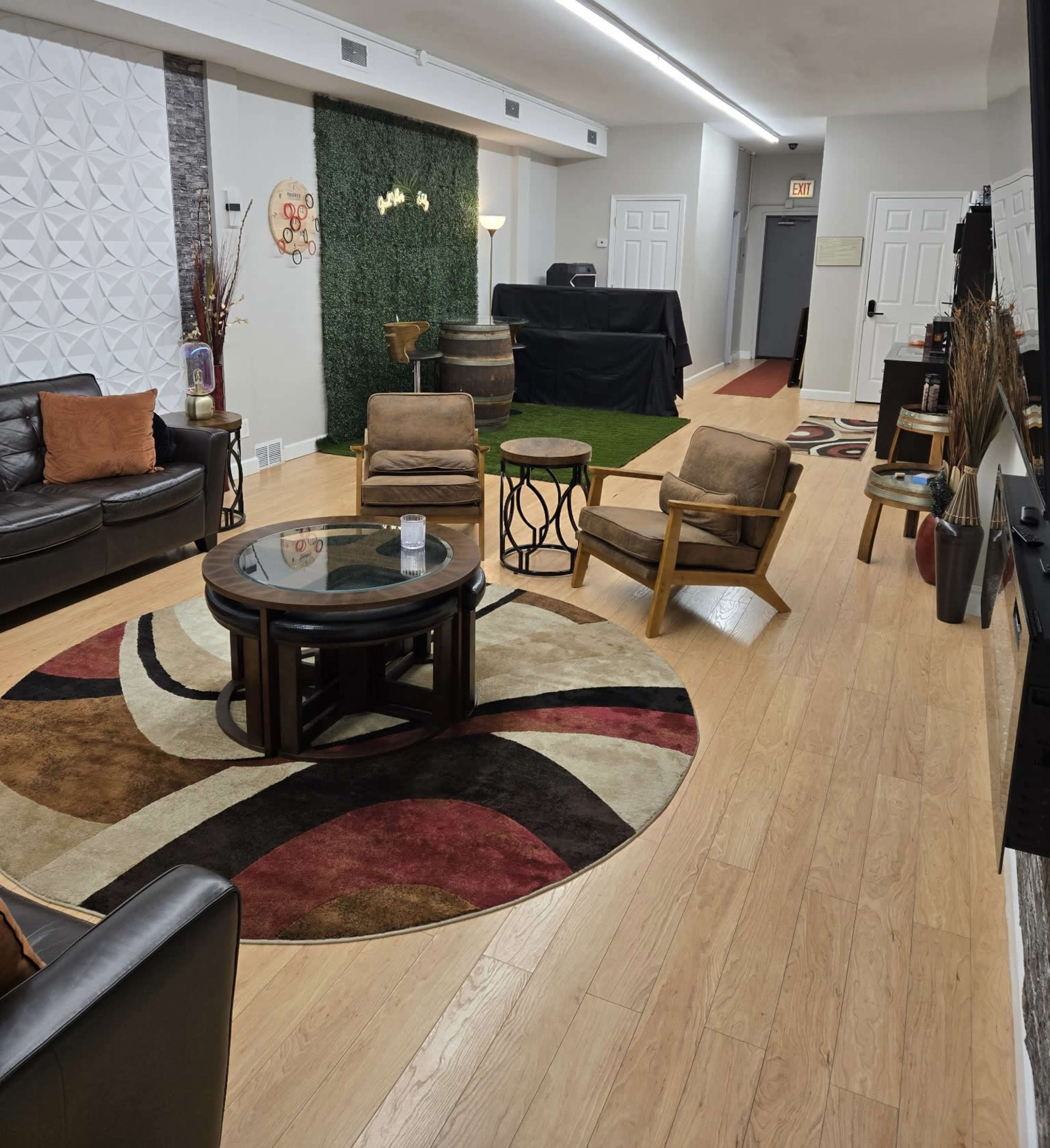 The image shows a cozy interior space featuring a seating area with leather couches and wooden chairs, a round coffee table, and a decorative rug, with a green wall and a barrel accent in the background.