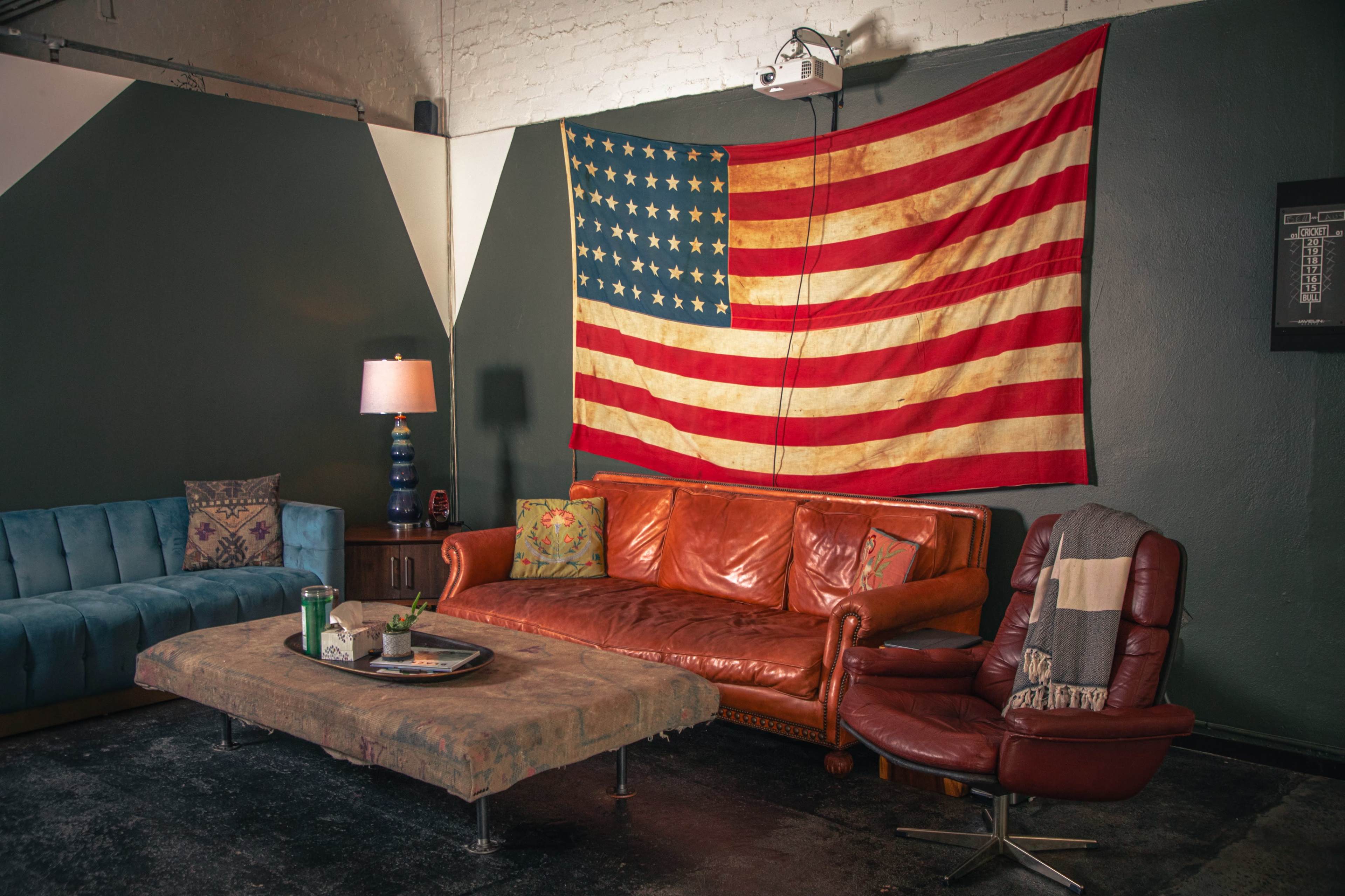 The image shows a cozy living space with a vintage leather sofa, a blue upholstered couch, a coffee table, and an American flag hanging on the wall.