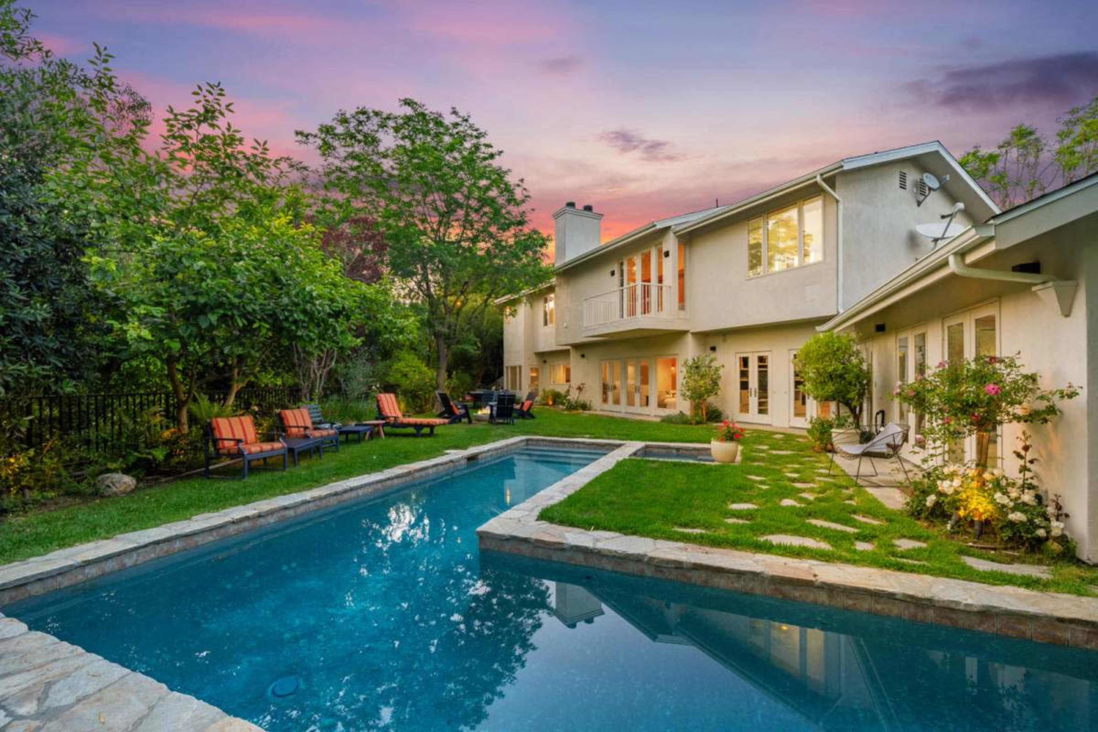 The image shows a backyard scene with a swimming pool, well-maintained grass, lounge chairs, and a two-story house in the background at dusk.