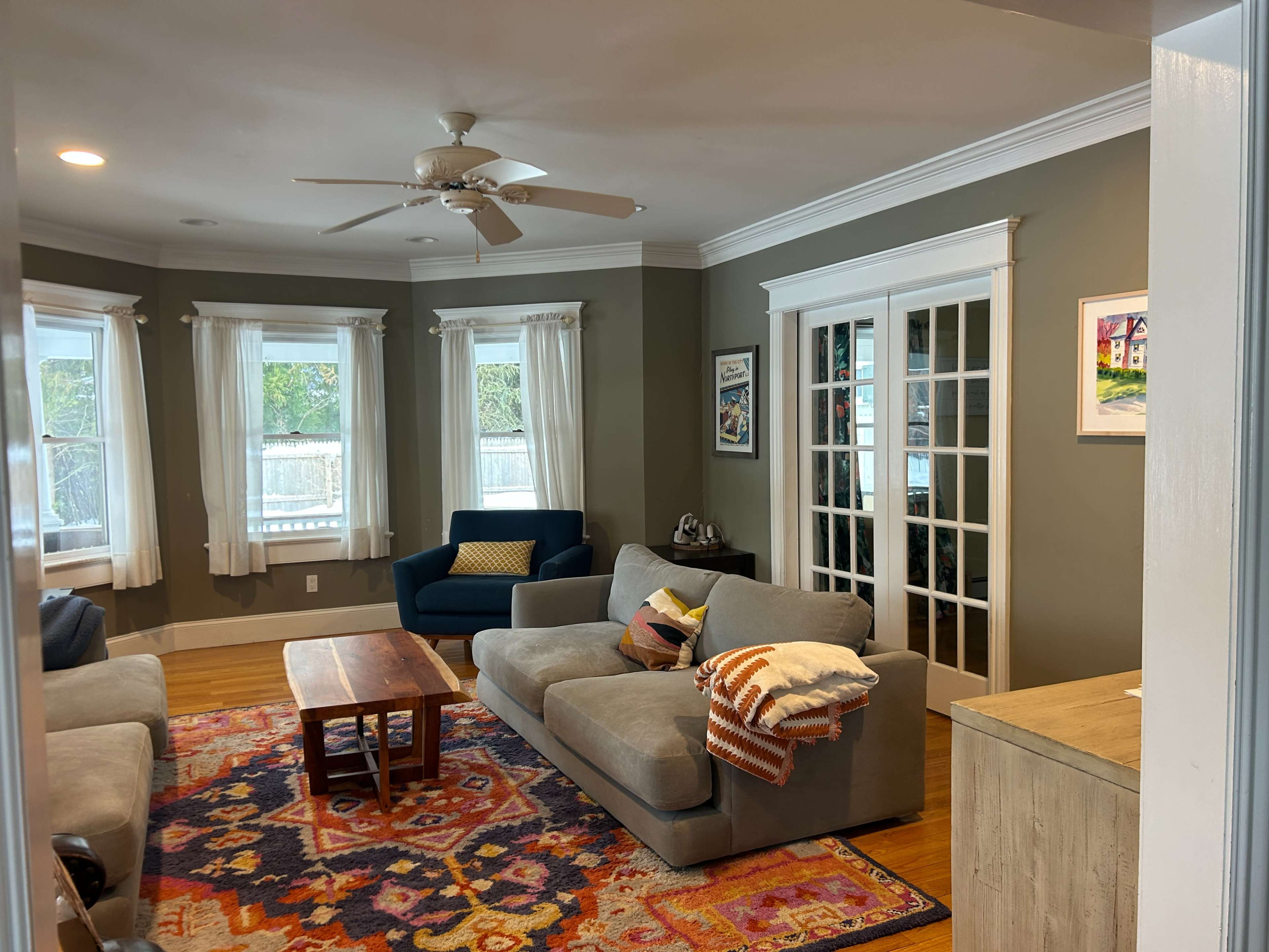 The image shows a cozy living room with a patterned rug, two sofas, a coffee table, and large windows adorned with white curtains.